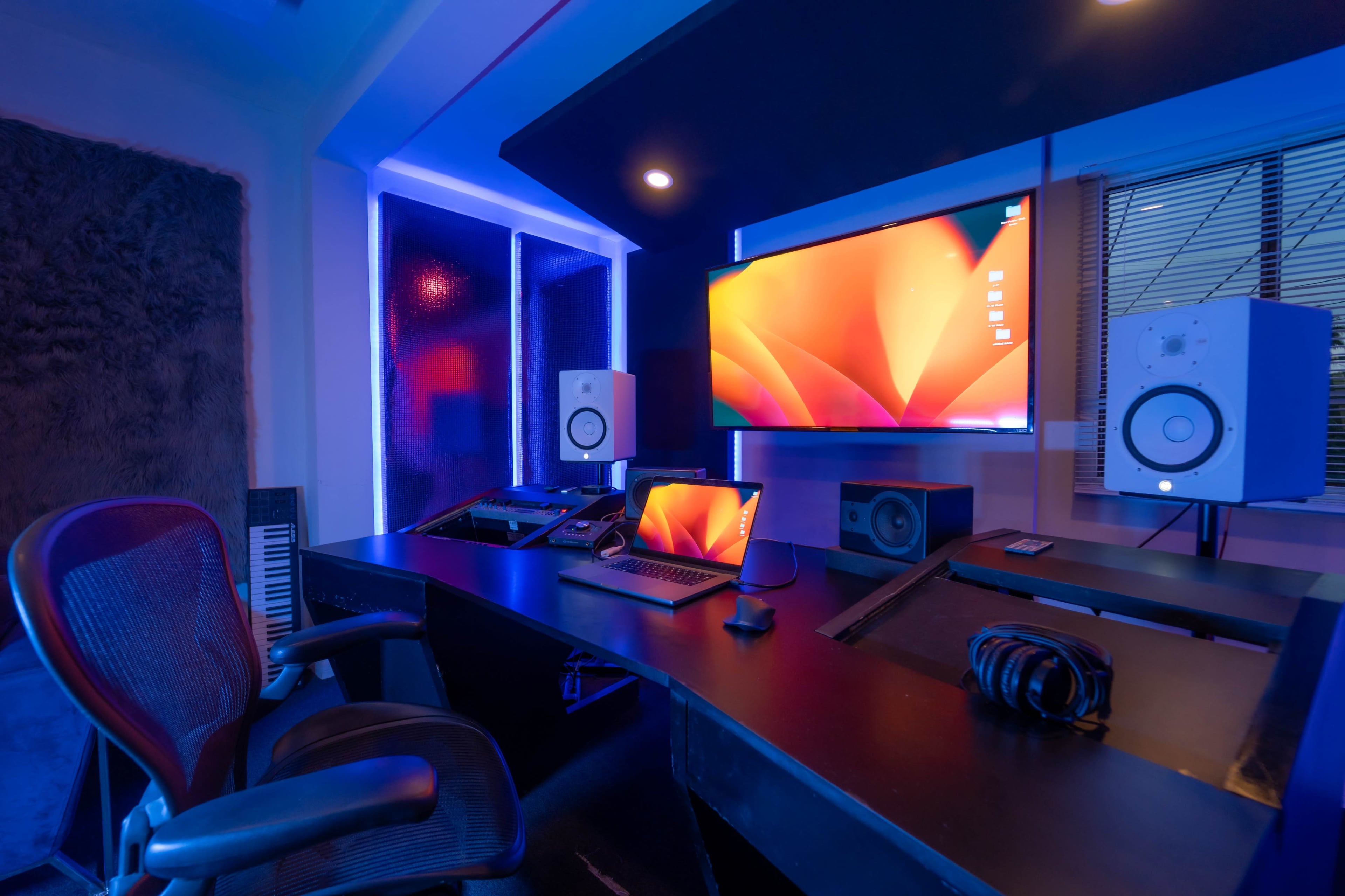 The image shows a modern music production studio featuring a desk with a laptop, audio monitors, and ambient blue lighting.