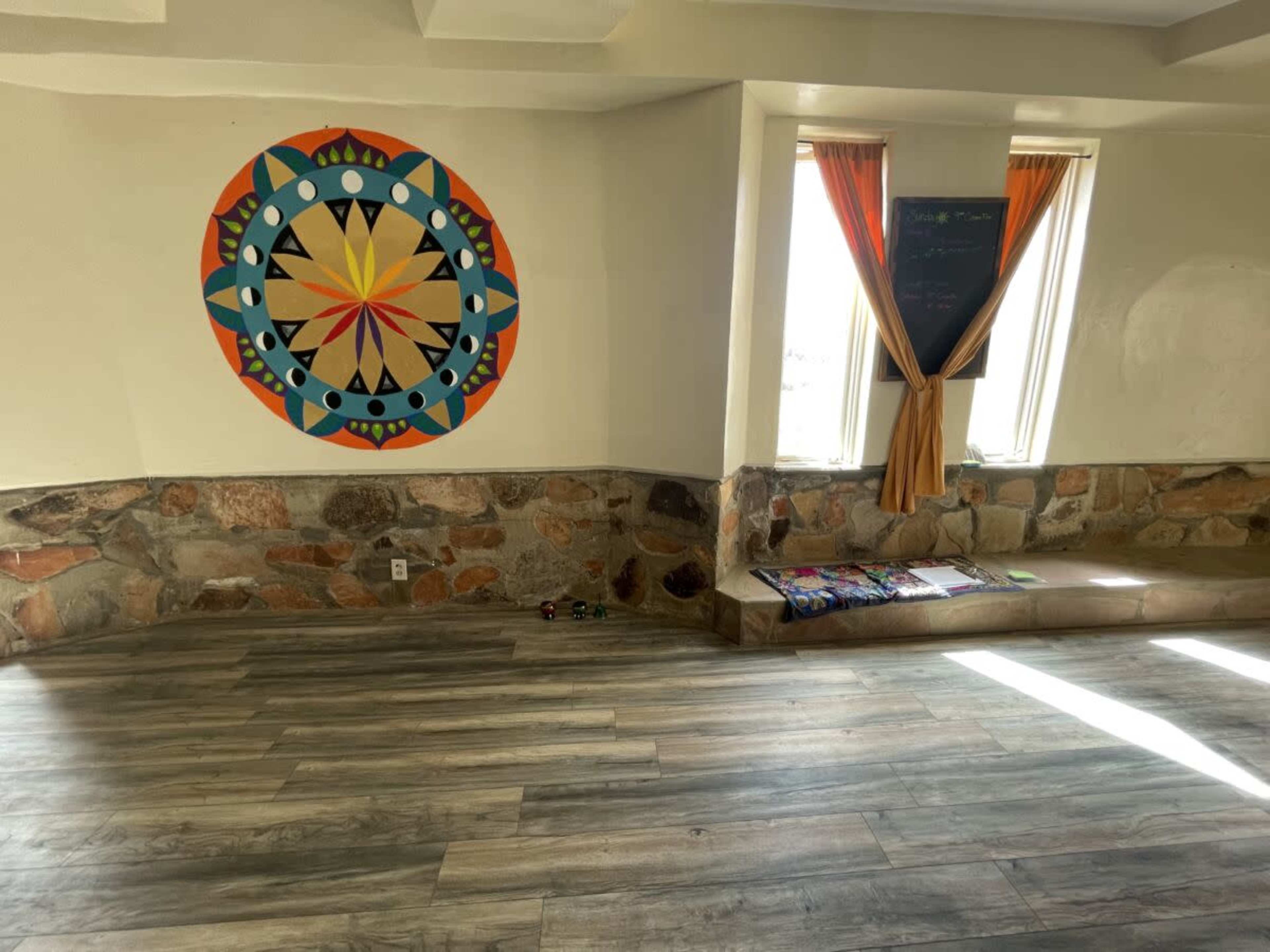 The image shows an interior room with a colorful mandala mural on one wall, large windows with orange curtains, and a stone accent at the base.