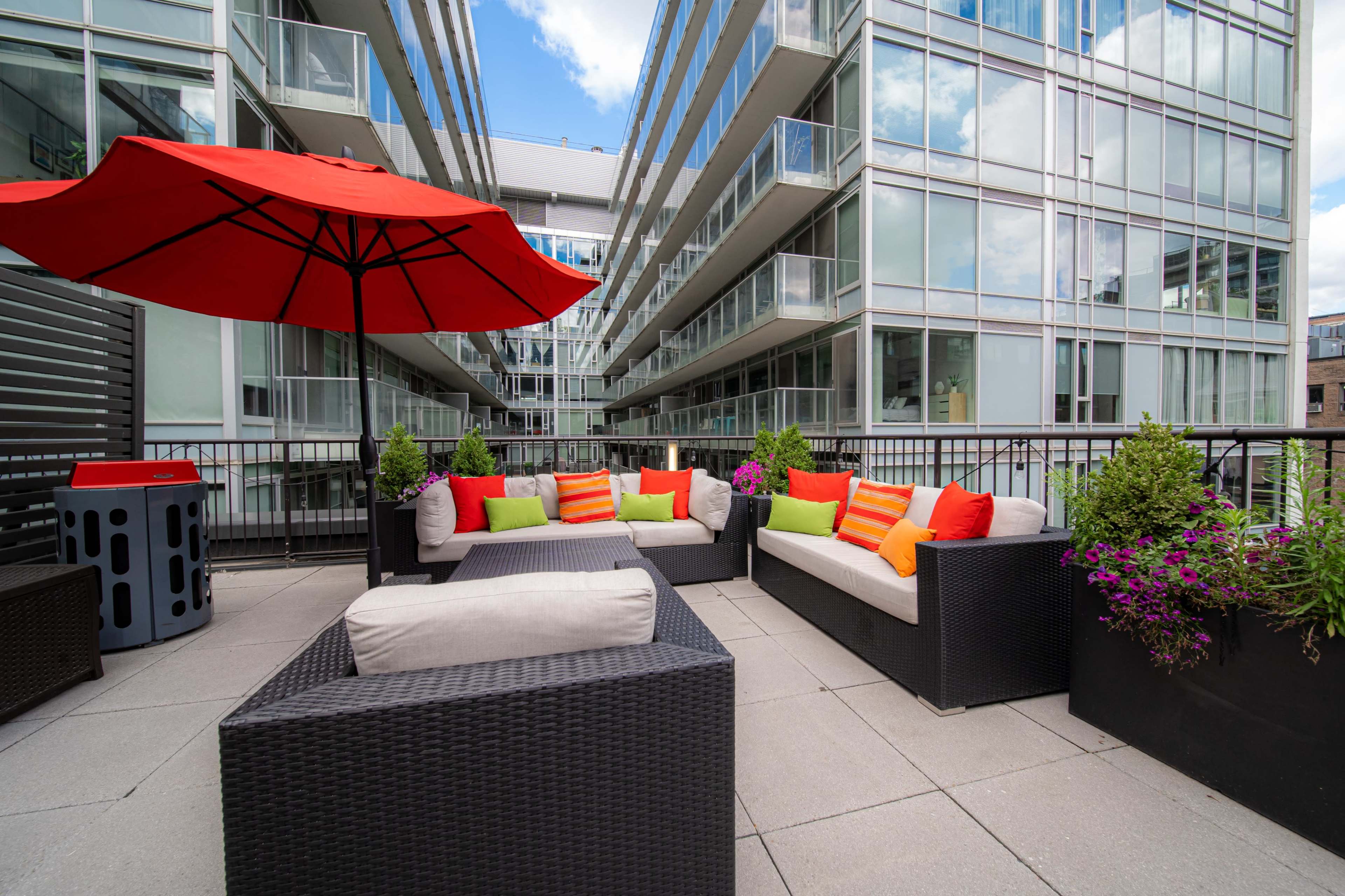 The image showcases a modern outdoor seating area with gray tiled flooring, multiple sofas adorned with colorful cushions, and a large red umbrella providing shade.