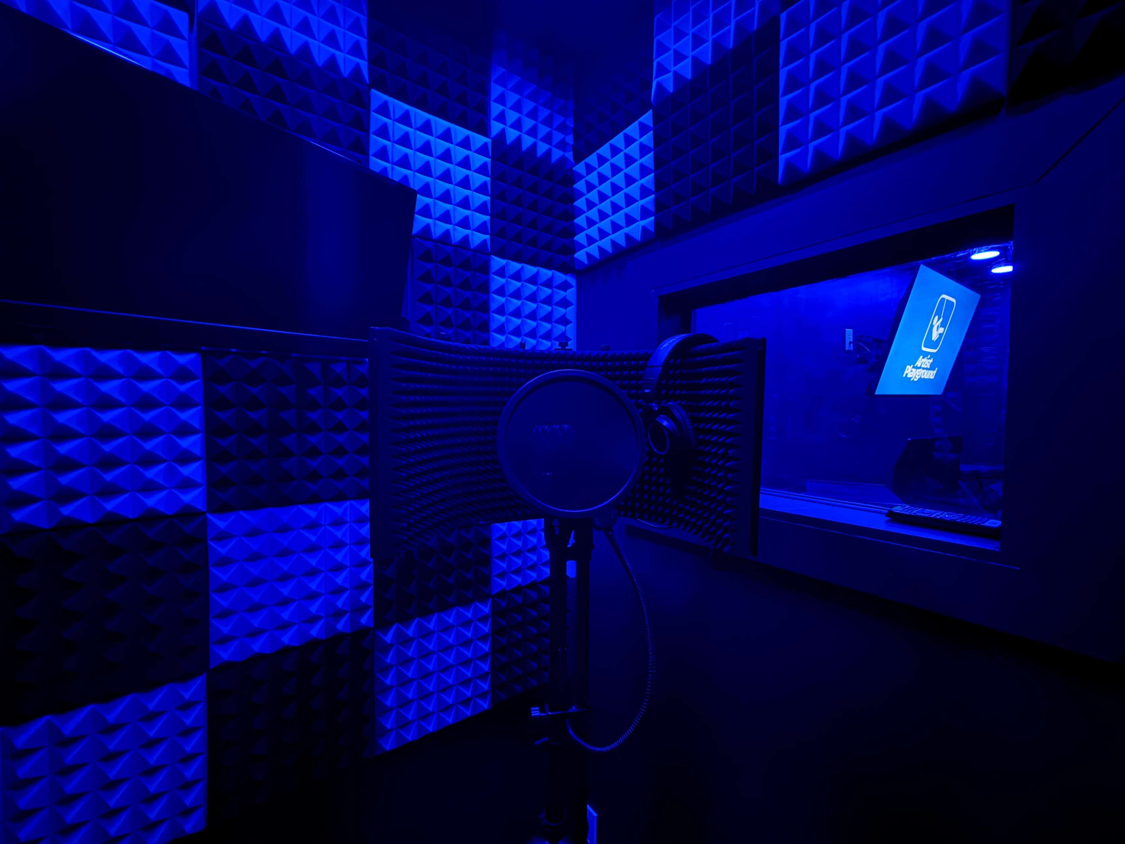 The image shows a recording studio booth with soundproof foam on the walls, illuminated by blue lighting, and a microphone set up in front of a glass window.