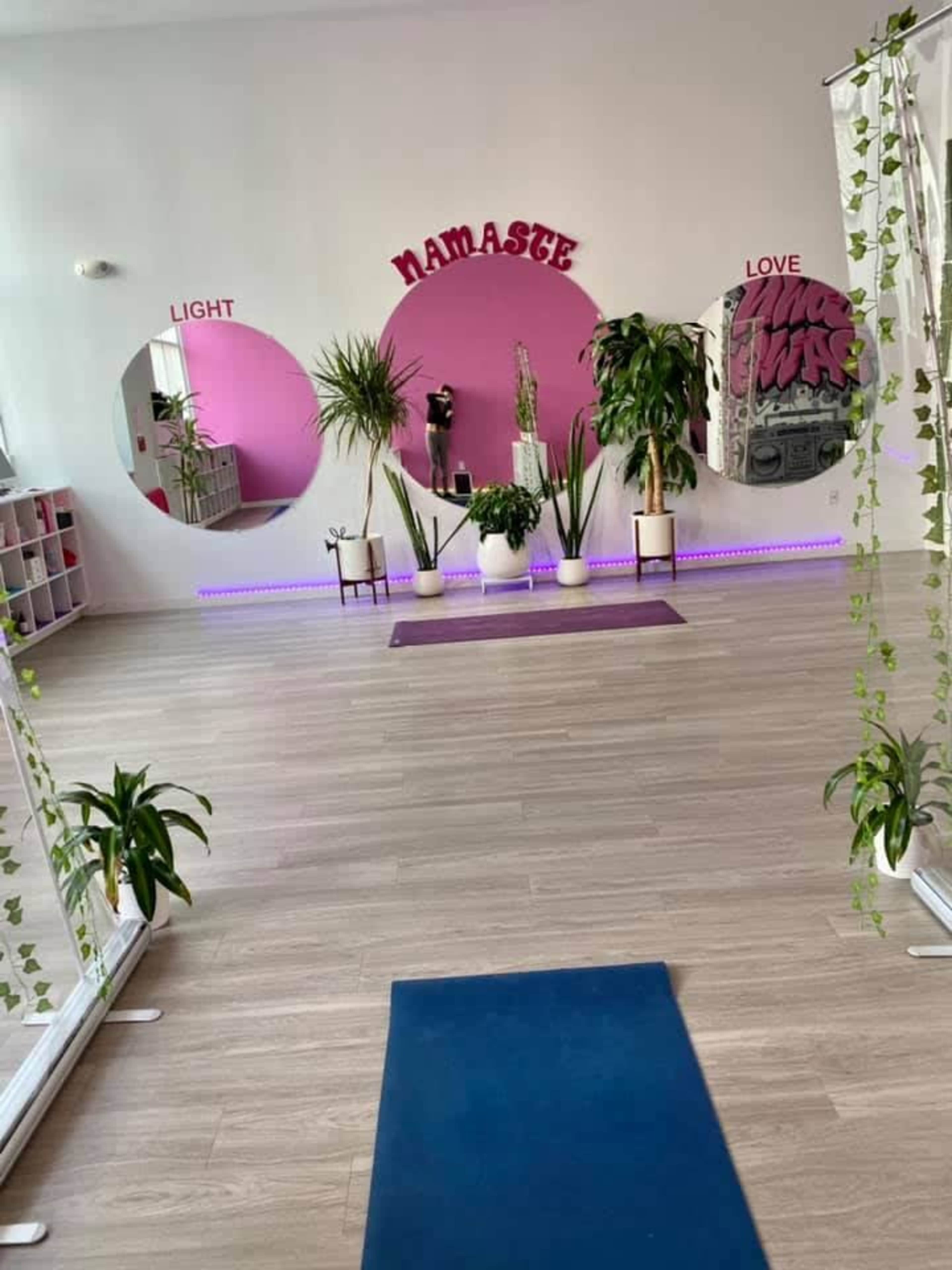 A brightly lit yoga studio featuring a pink wall with the words "NAMASTE," "LIGHT," and "LOVE," surrounded by plants and large circular mirrors.