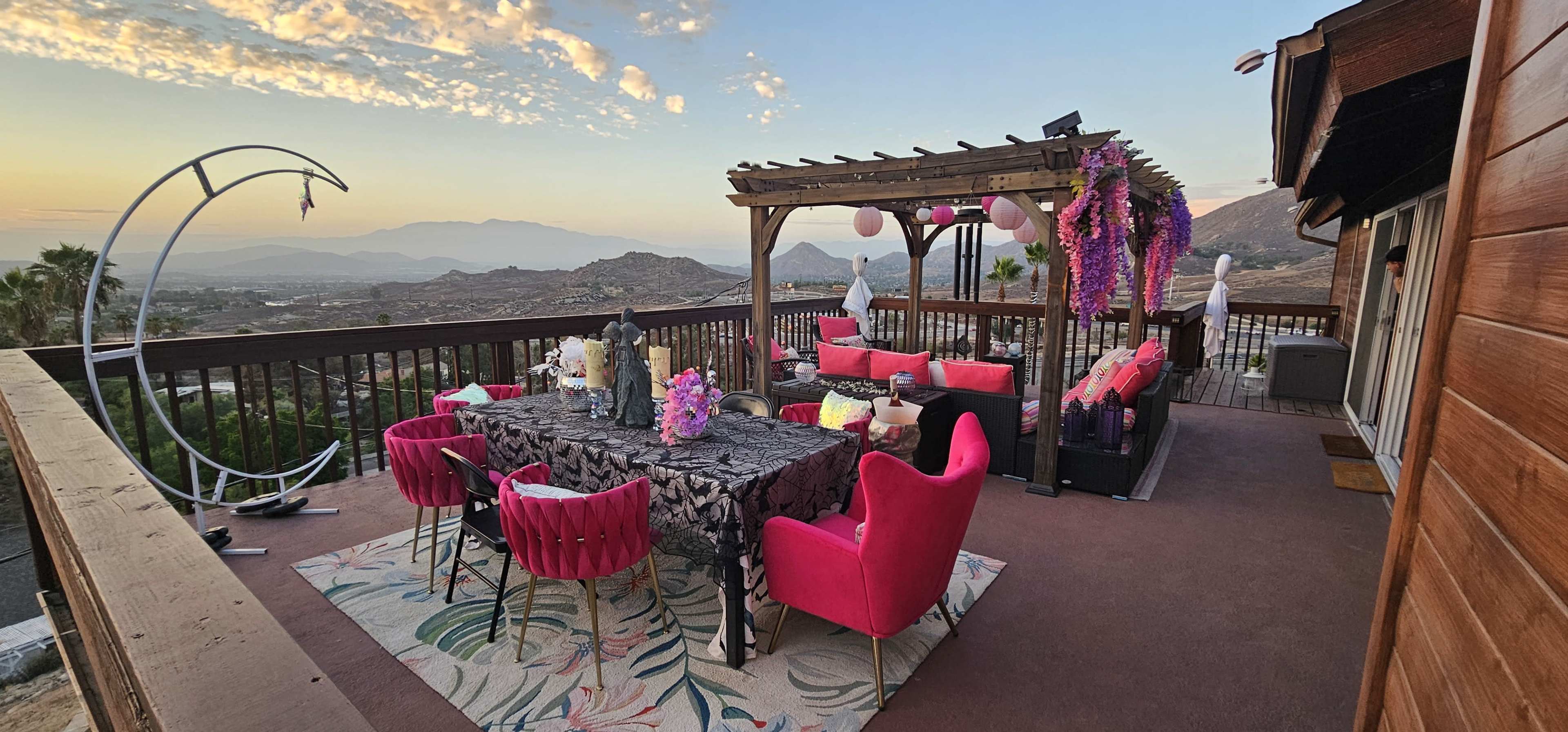 A spacious outdoor patio is adorned with pink furniture, a decorative table, and hanging flowers, set against a backdrop of mountains and a sunset sky.