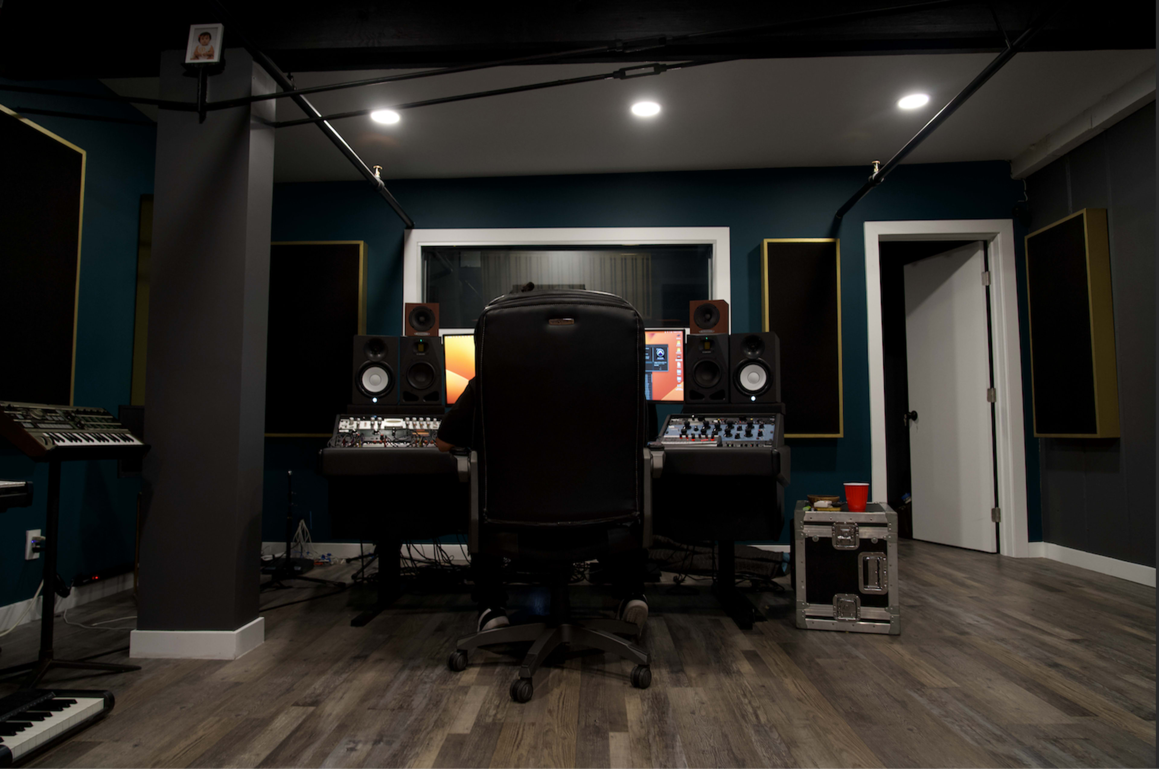 A person sits at a recording studio desk with audio equipment and speakers, facing a computer screen in a dimly lit room.