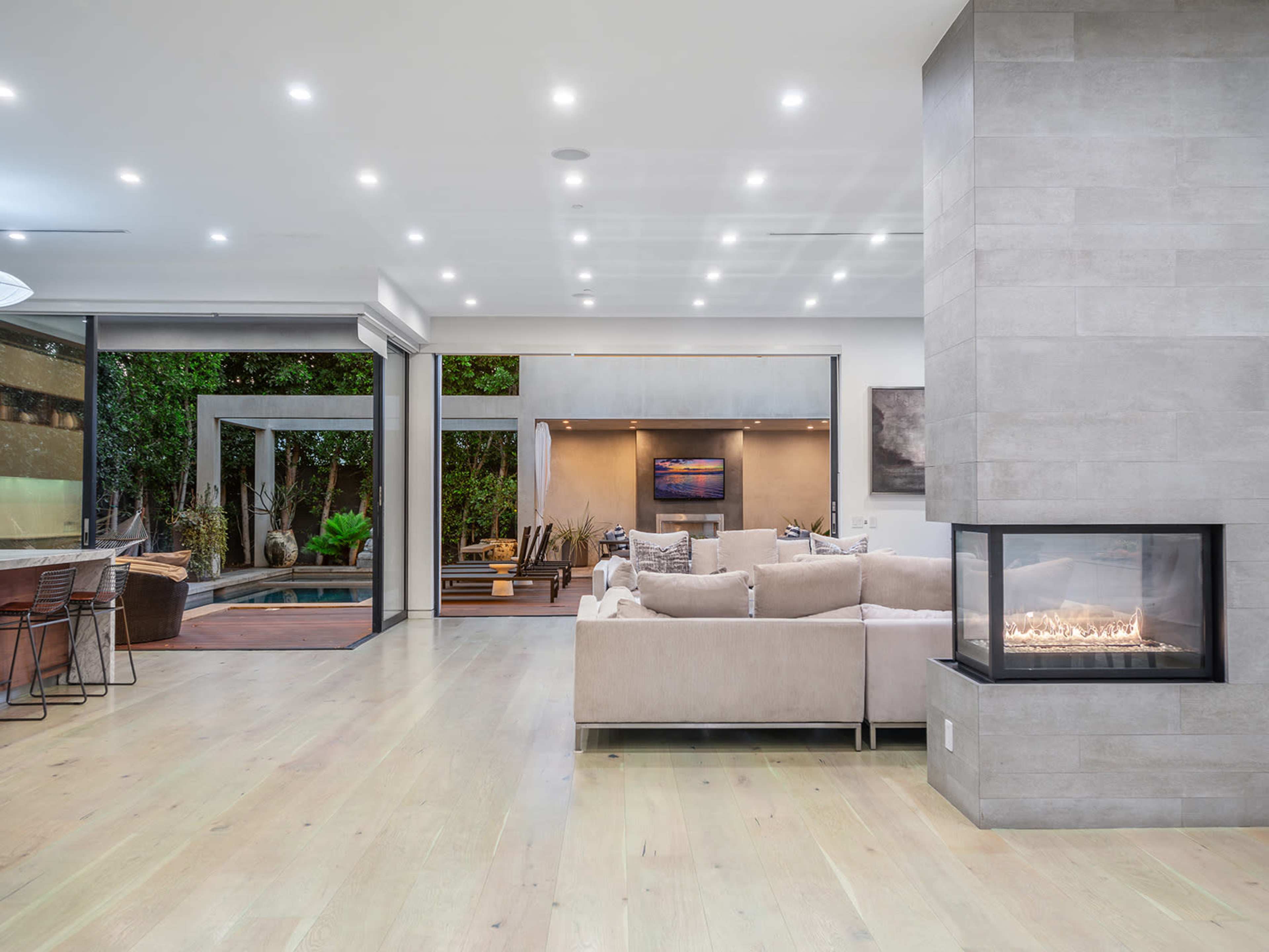 The image shows a modern living room with a fireplace, large windows, and access to an outdoor area featuring greenery and seating.