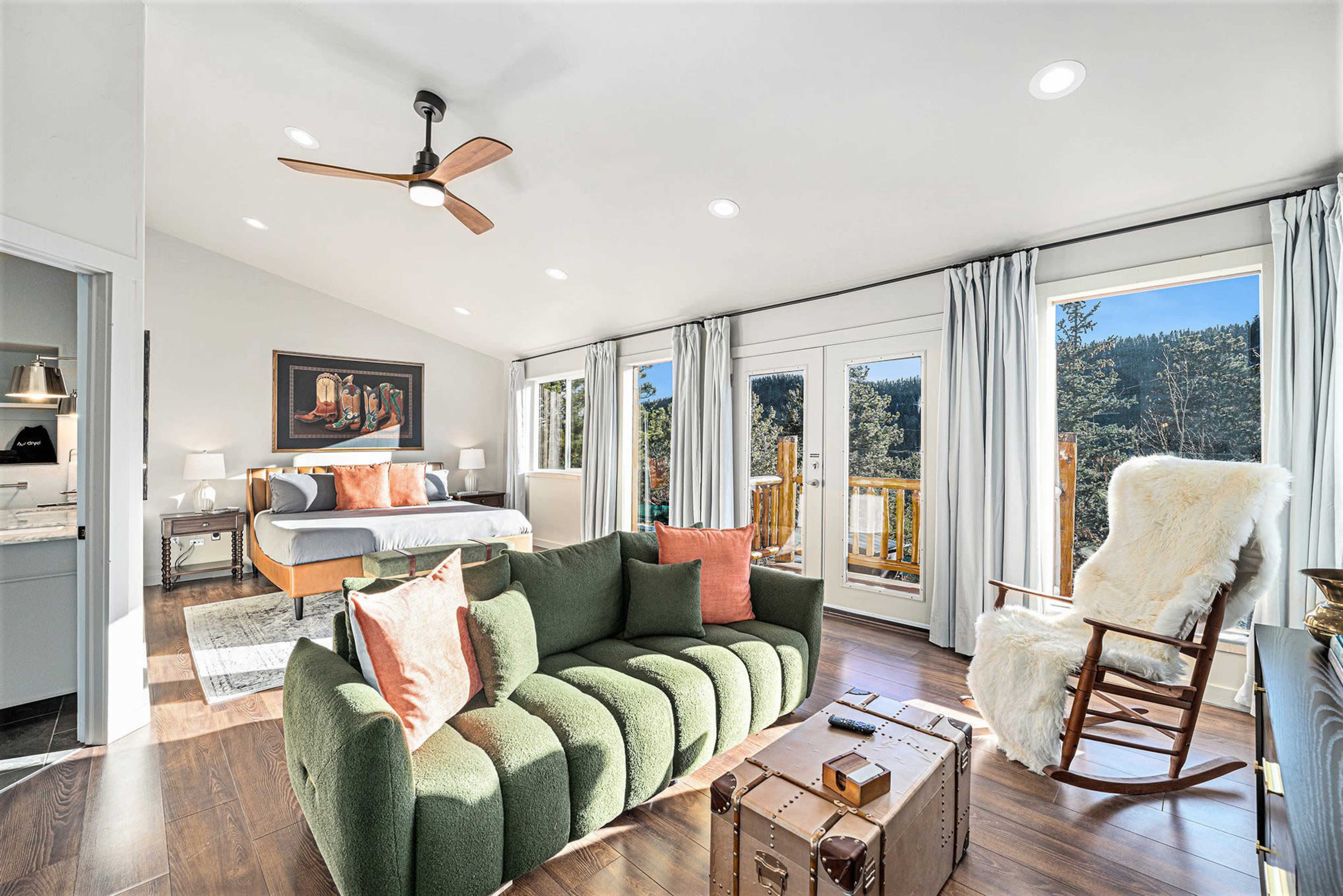 The image shows a modern bedroom with a large bed, a green sofa, a coffee table, and large windows overlooking a forested area.