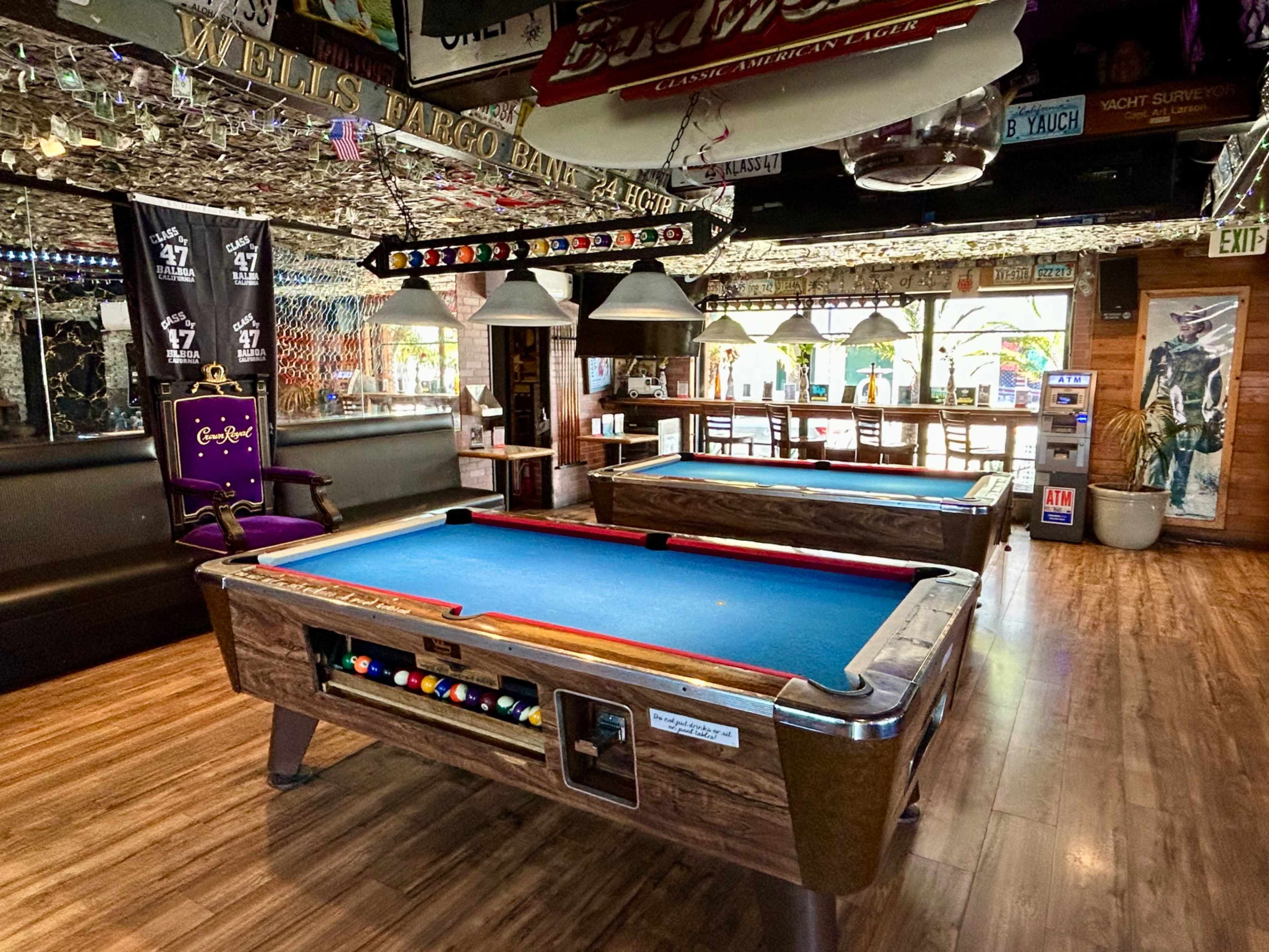 The image shows a spacious, well-lit bar area featuring two pool tables, surrounded by seating and decorated with various signs and memorabilia.