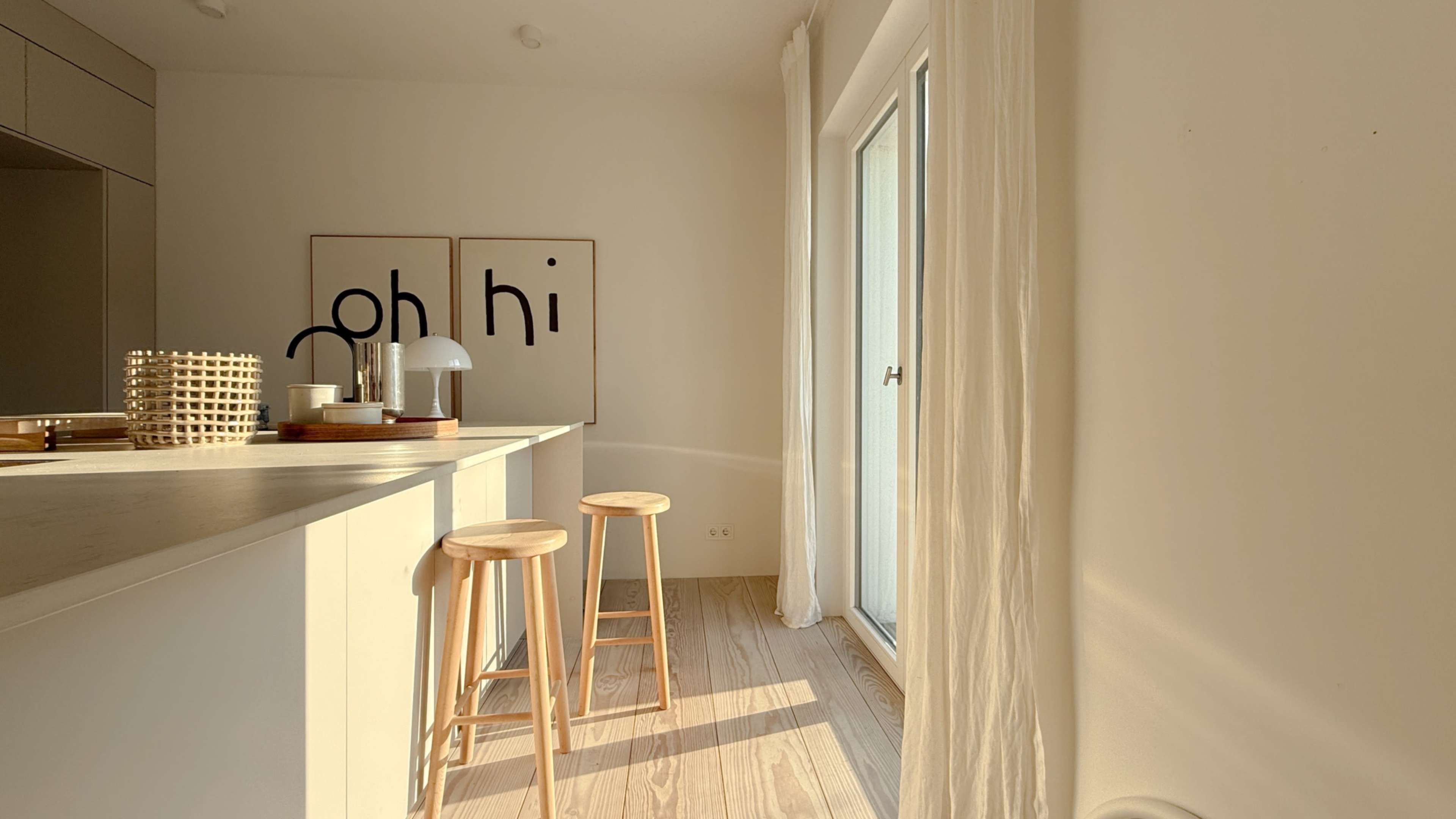 A bright, minimalist kitchen features a countertop with two wooden stools and framed prints on the wall.