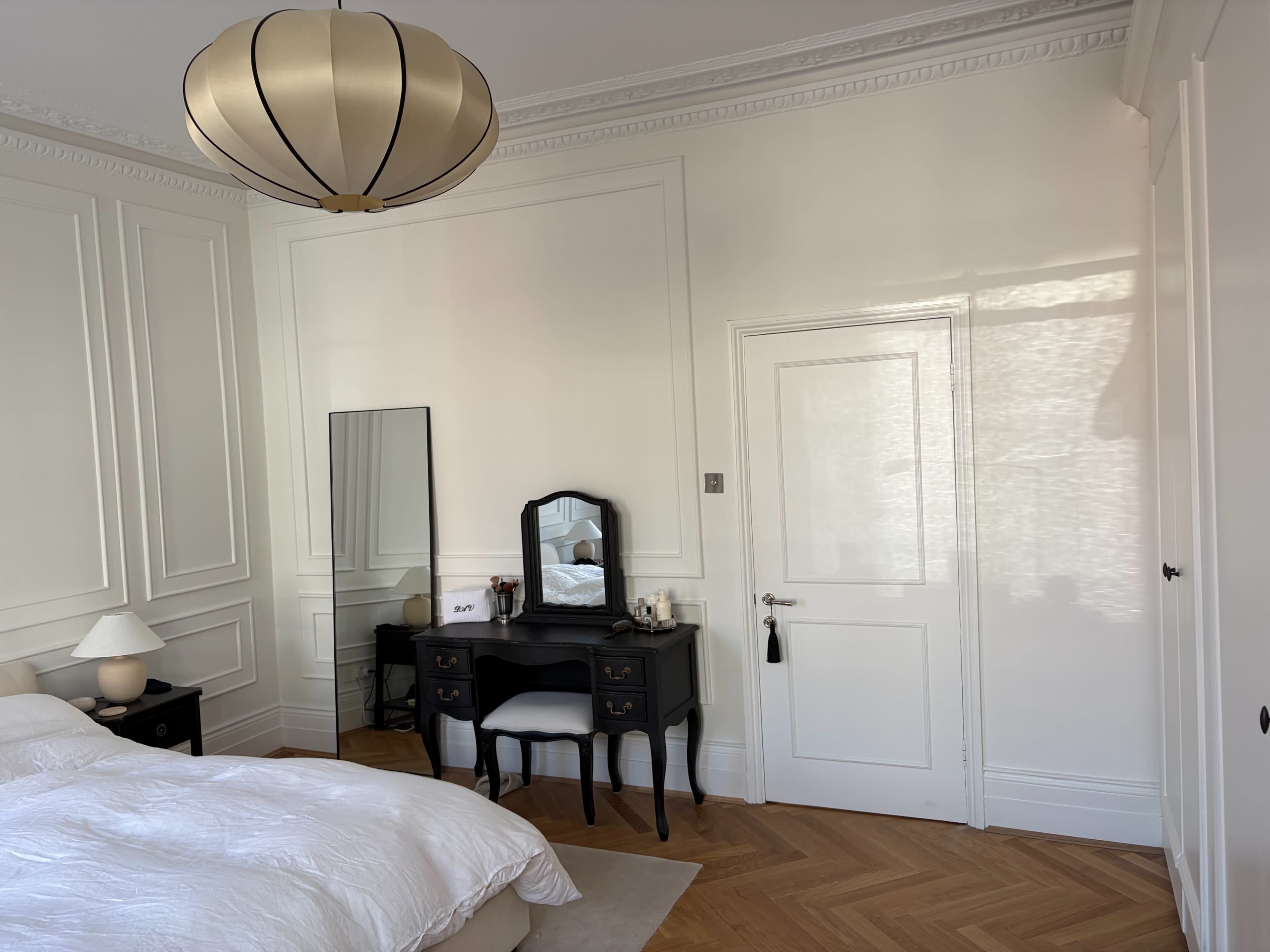 The image shows a neatly arranged bedroom with a large bed, a black vanity table with a mirror, and a light fixture hanging from the ceiling.