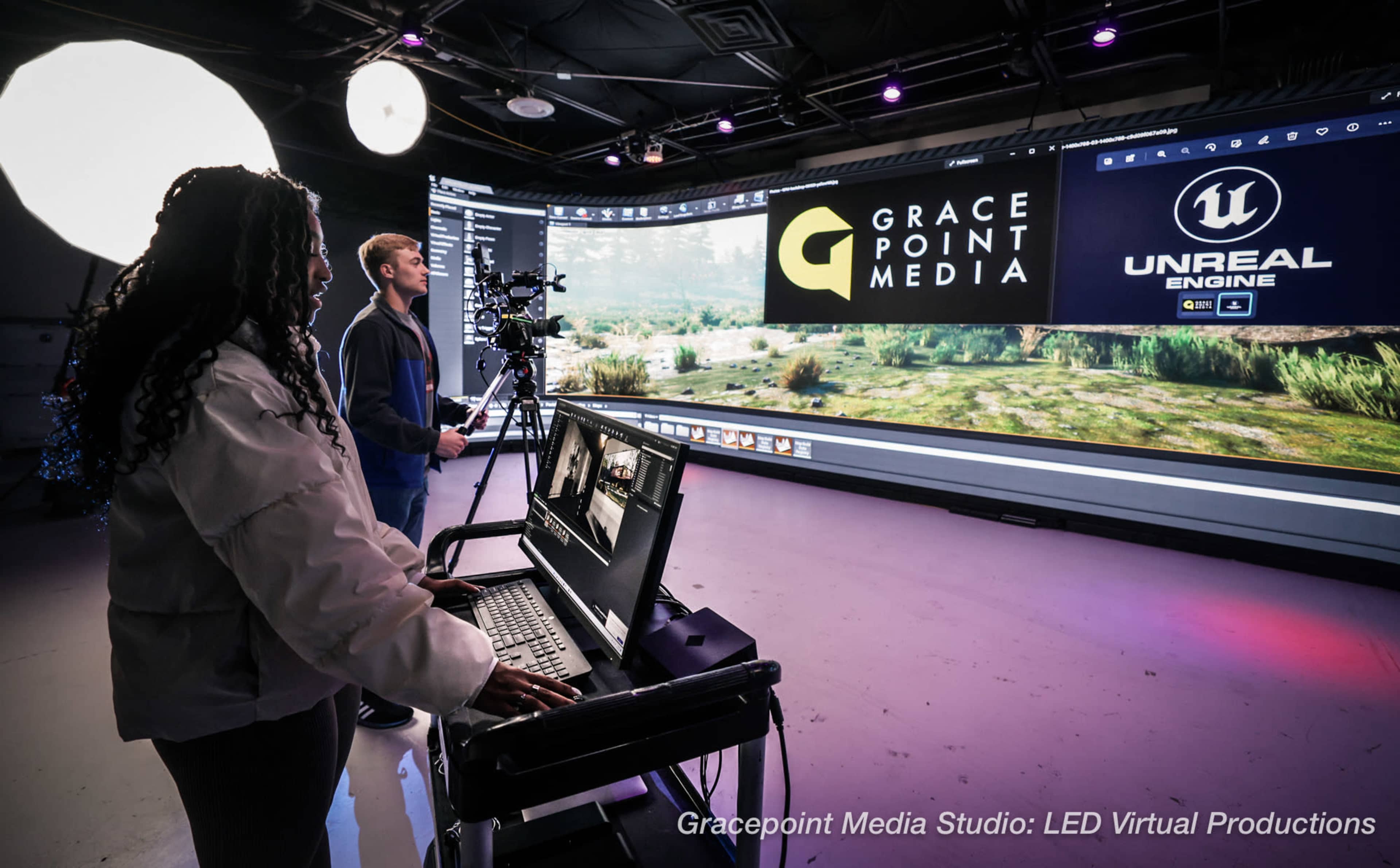 A woman operates a computer while a man sets up a camera in a studio featuring a large LED screen displaying a virtual environment.