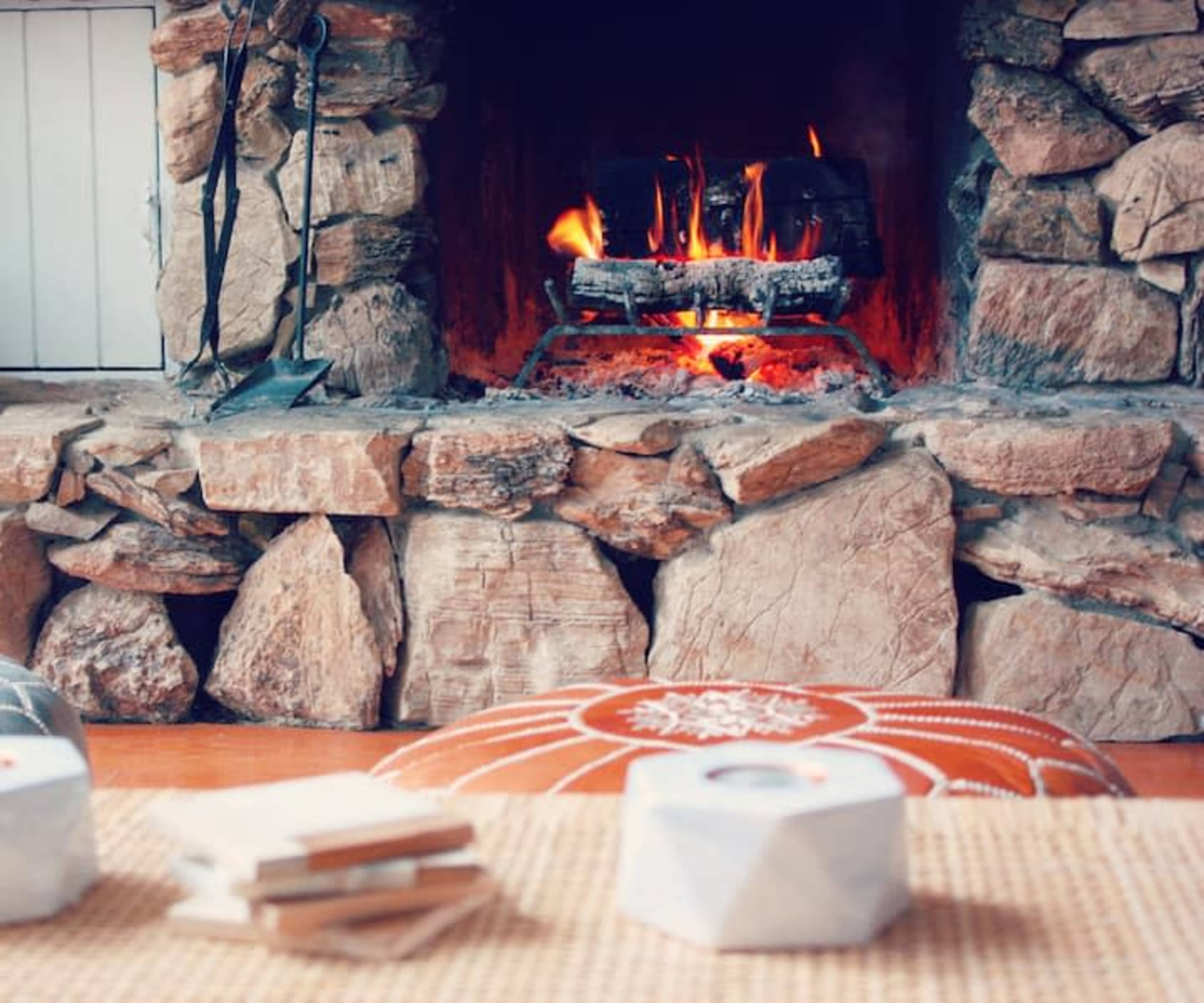 A stone fireplace with a roaring fire is the focal point of a cozy interior, complemented by decorative cushions and a small stack of coasters in the foreground.