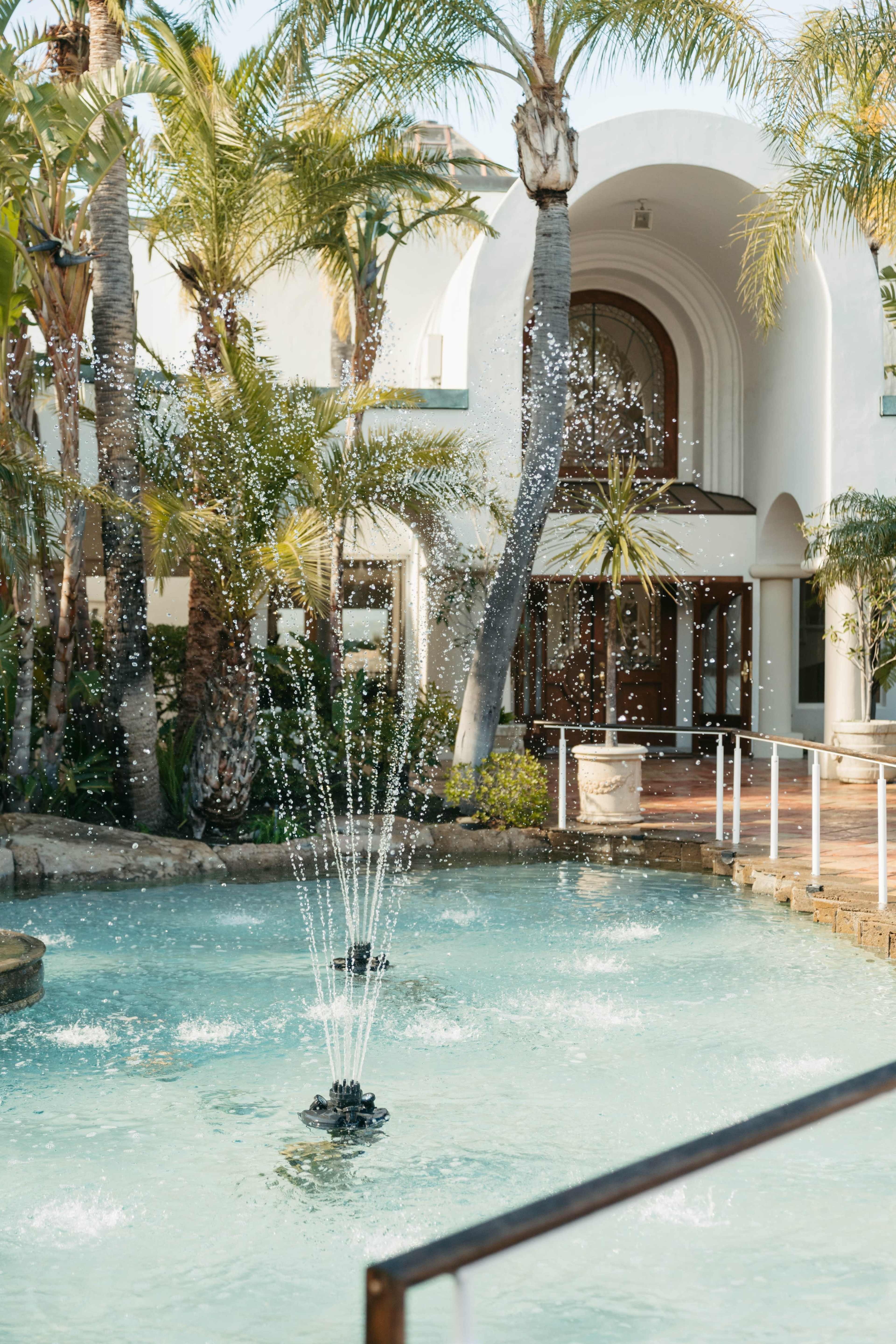A fountain sprays water in a clear pool surrounded by palm trees and architecture featuring arched entrances.