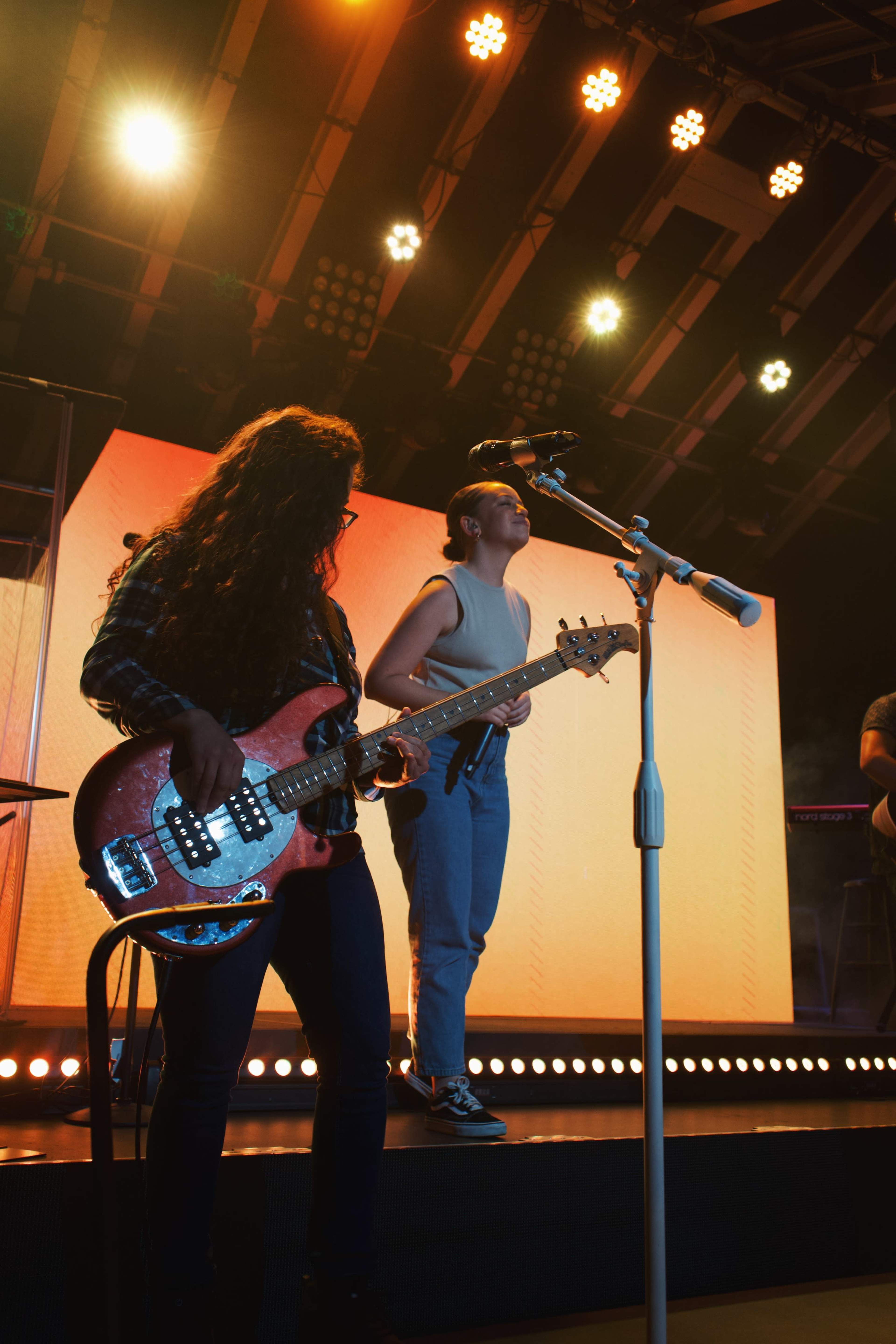 A musician plays a bass guitar while another vocalist performs on stage with colorful lighting in the background.
