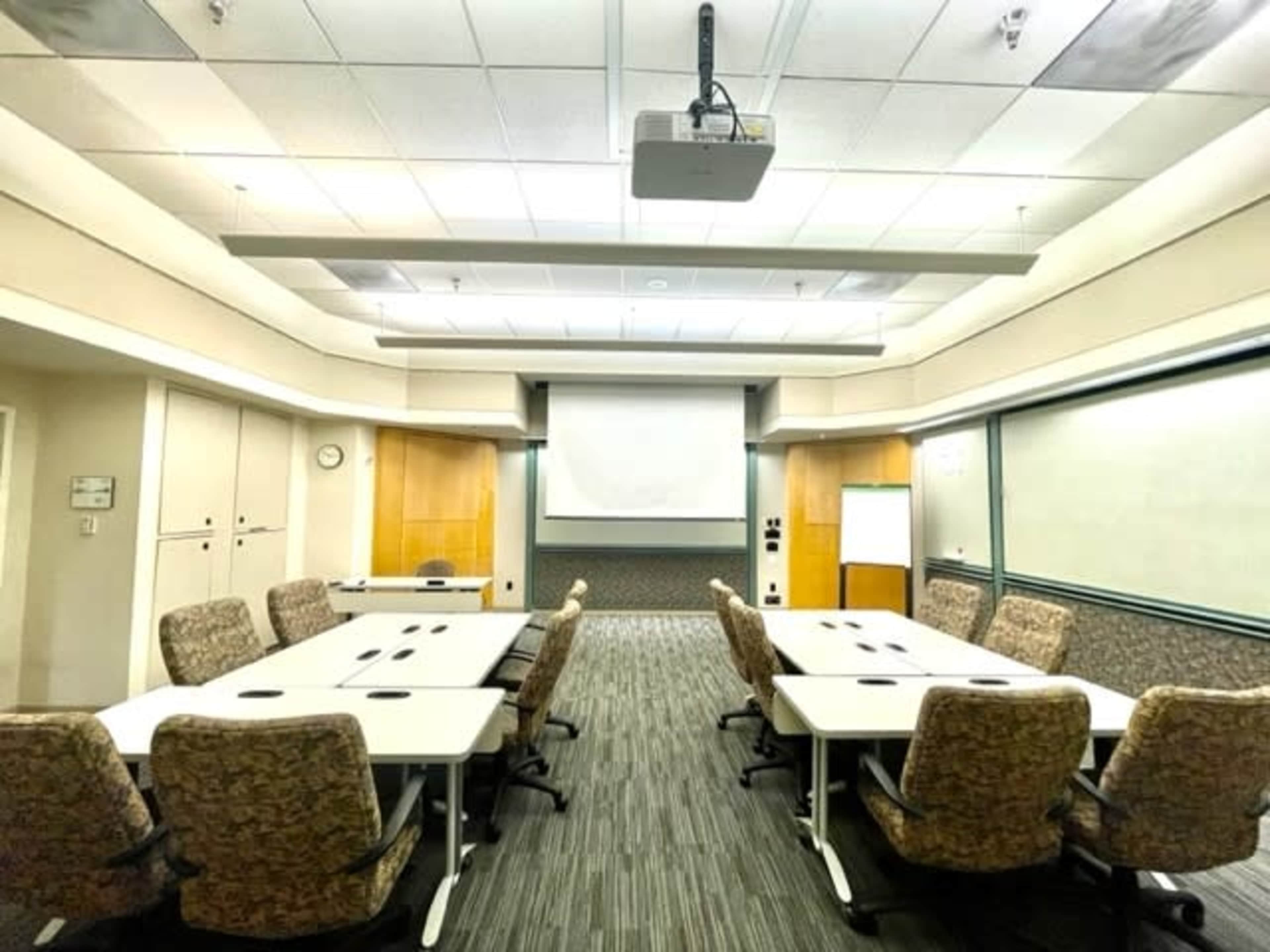 A conference room is set up with several tables arranged in a U-shape and chairs along the sides, featuring a projector and a large screen at the front.