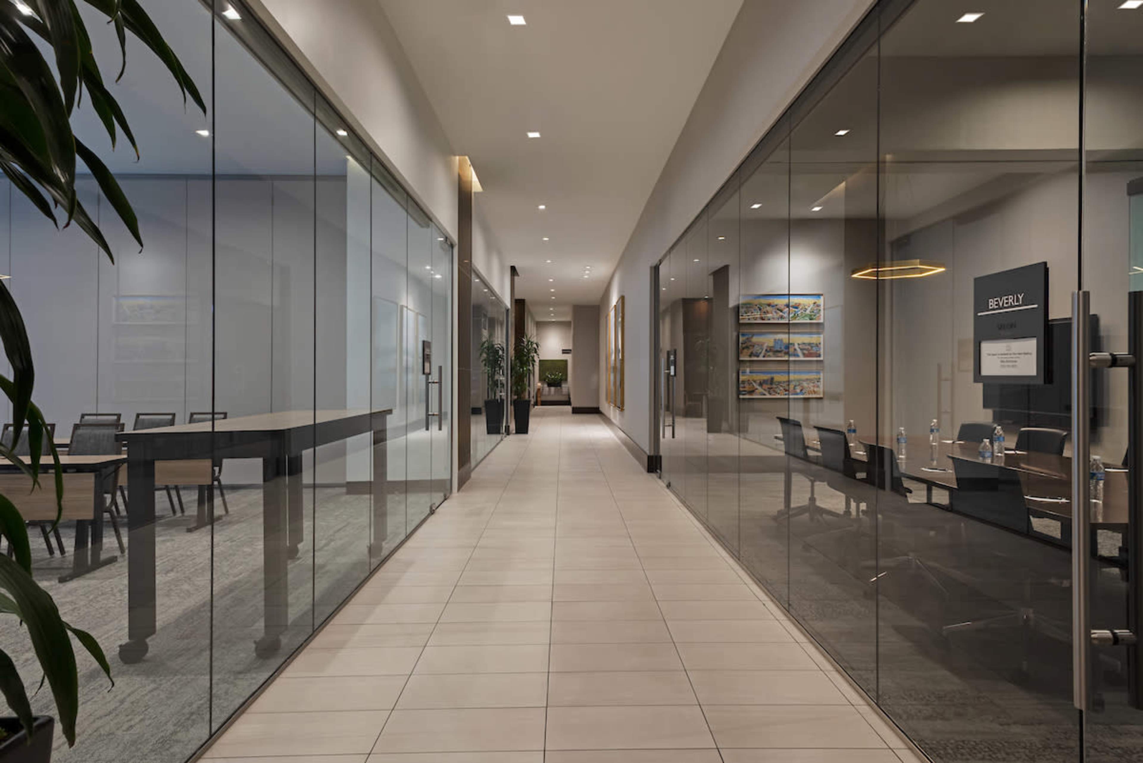 A long corridor features glass-walled meeting rooms and a conference area with a table, illuminated by overhead lights.