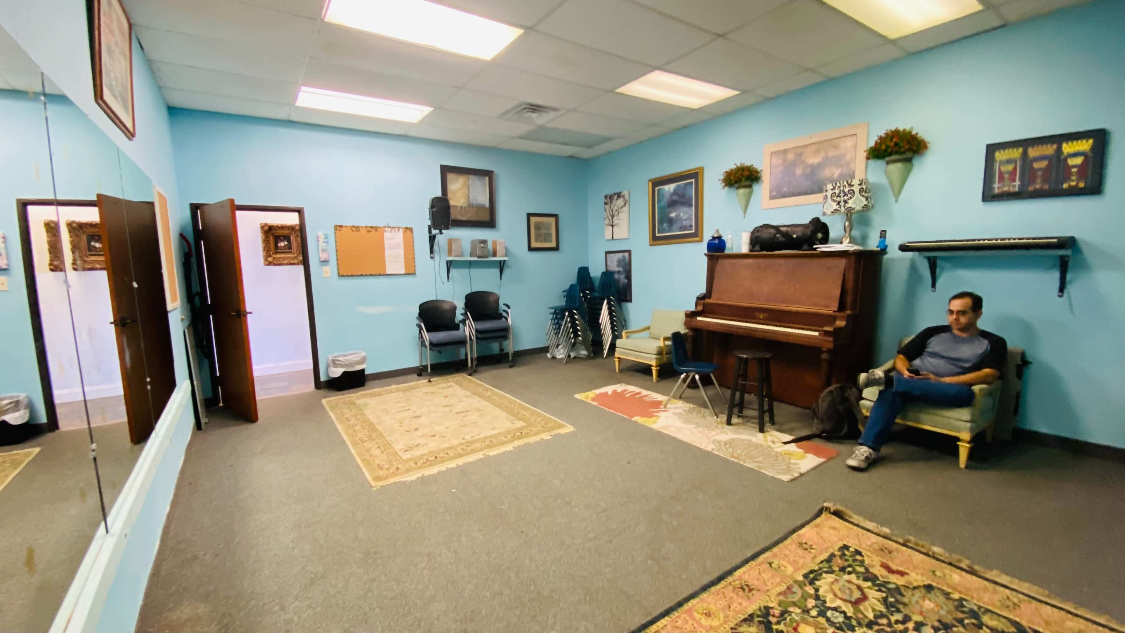 The image shows a light blue room with a piano, a guitar, chairs, and decorative rugs on the floor.