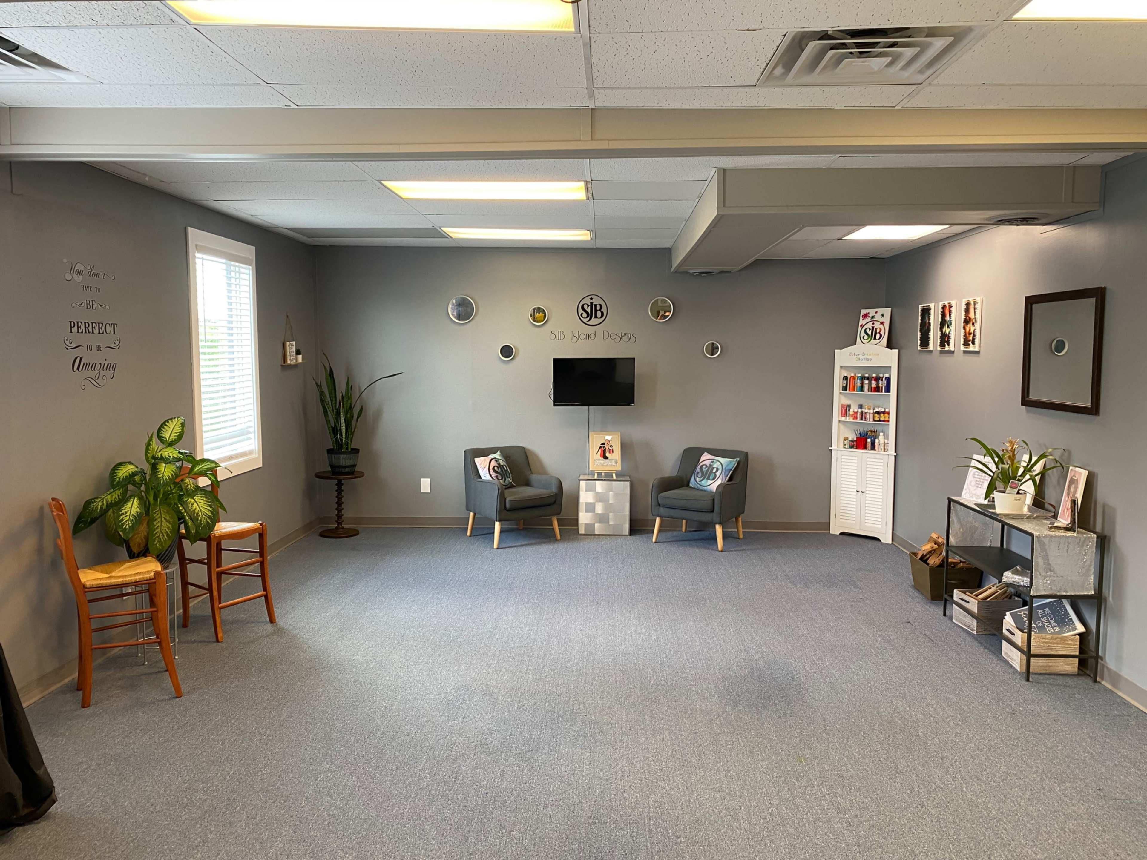 A spacious room with gray walls, two chairs, a small table, a television on the wall, and decorative items displayed on shelves.