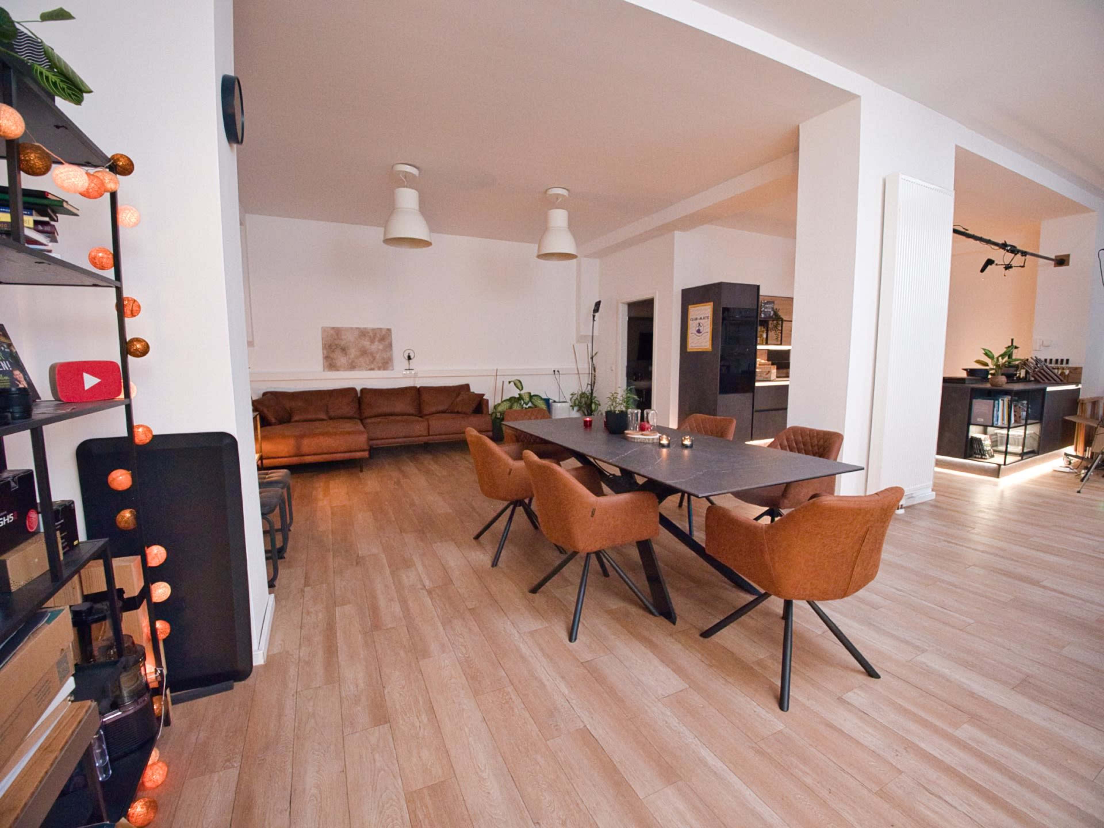 The image shows a spacious, modern interior with a large dining table surrounded by chairs, a brown sofa in the background, and wooden flooring.