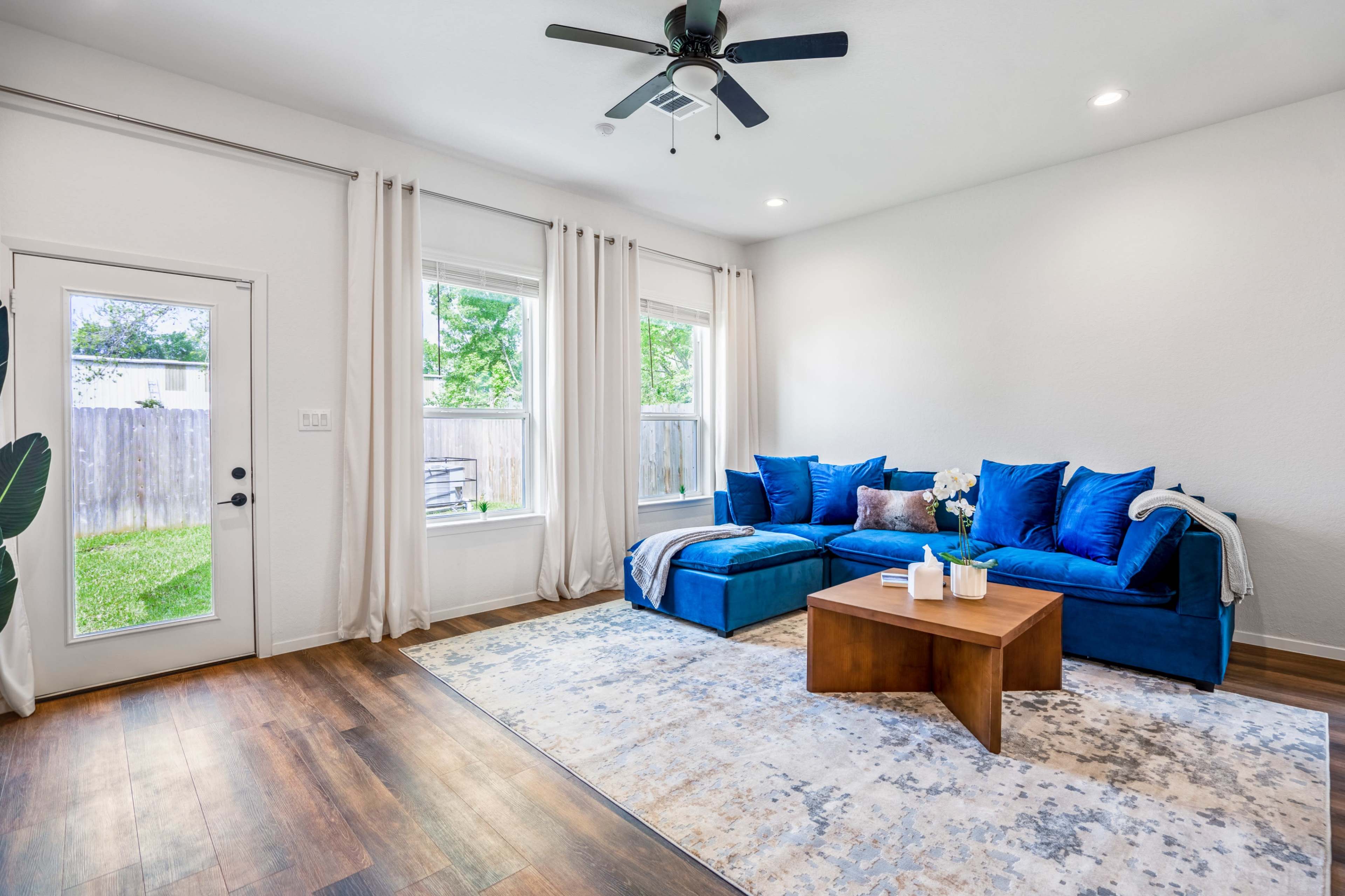 A bright living room features a blue sectional sofa, a wooden coffee table, and large windows with sheer curtains.