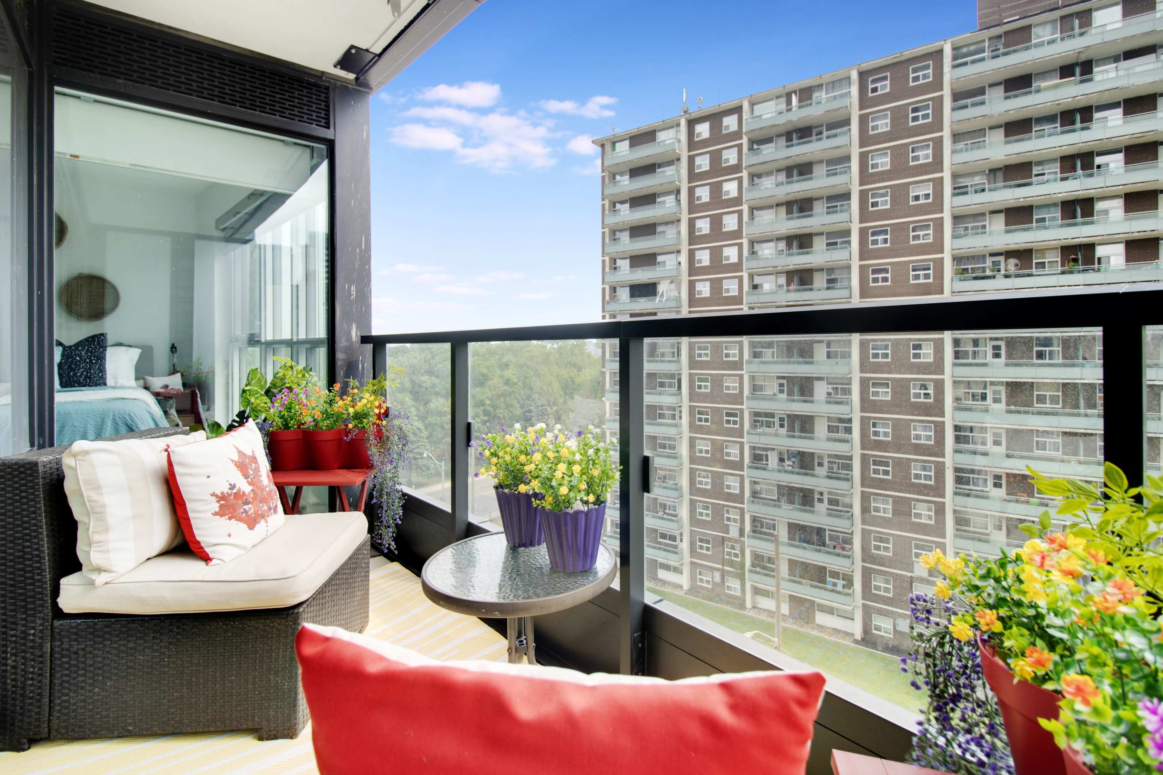 A balcony with potted plants and outdoor furniture overlooks an apartment building and a clear blue sky.