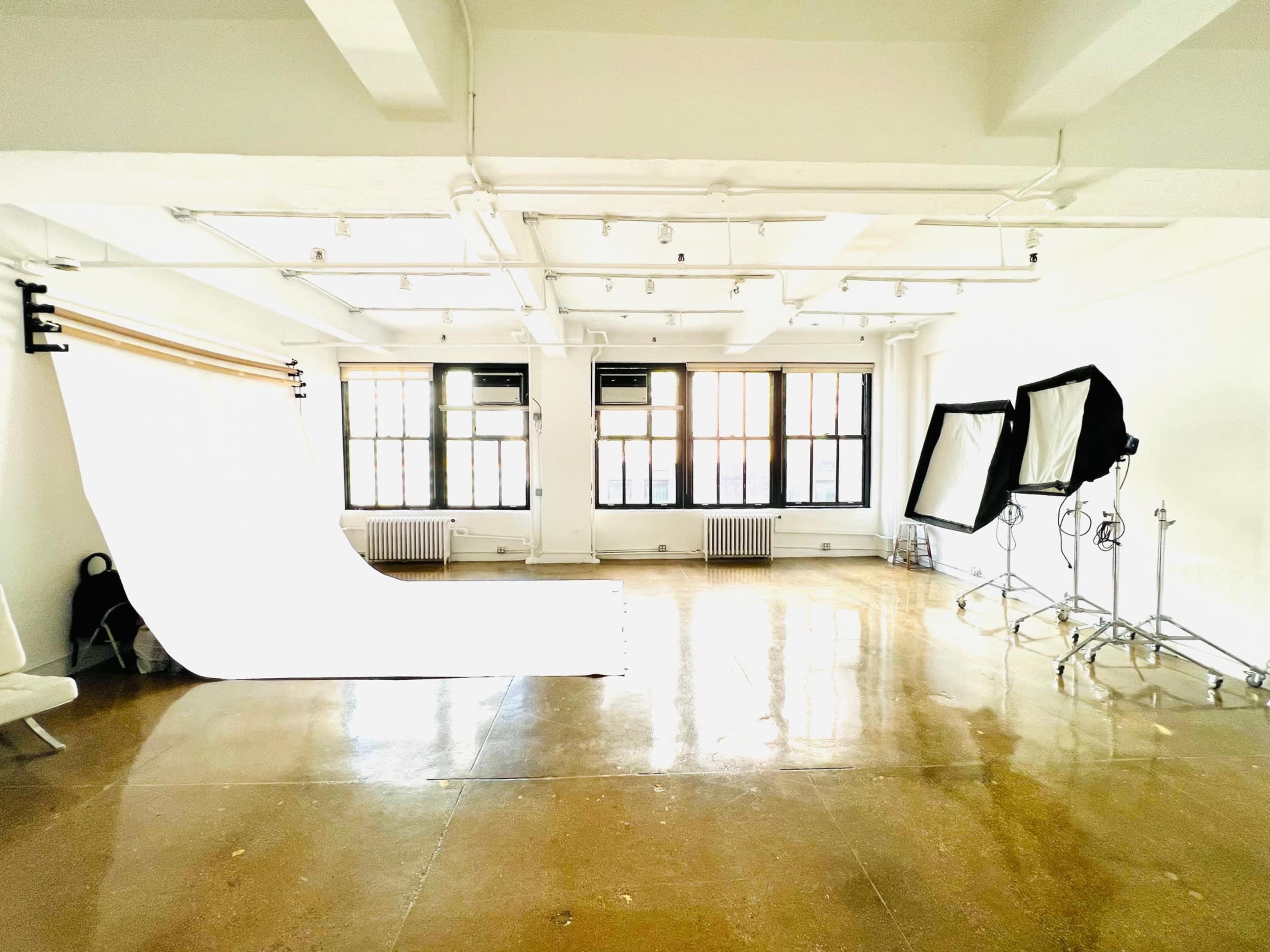 The image shows a bright photography studio with large windows, a seamless backdrop, and lighting equipment arranged on the floor.