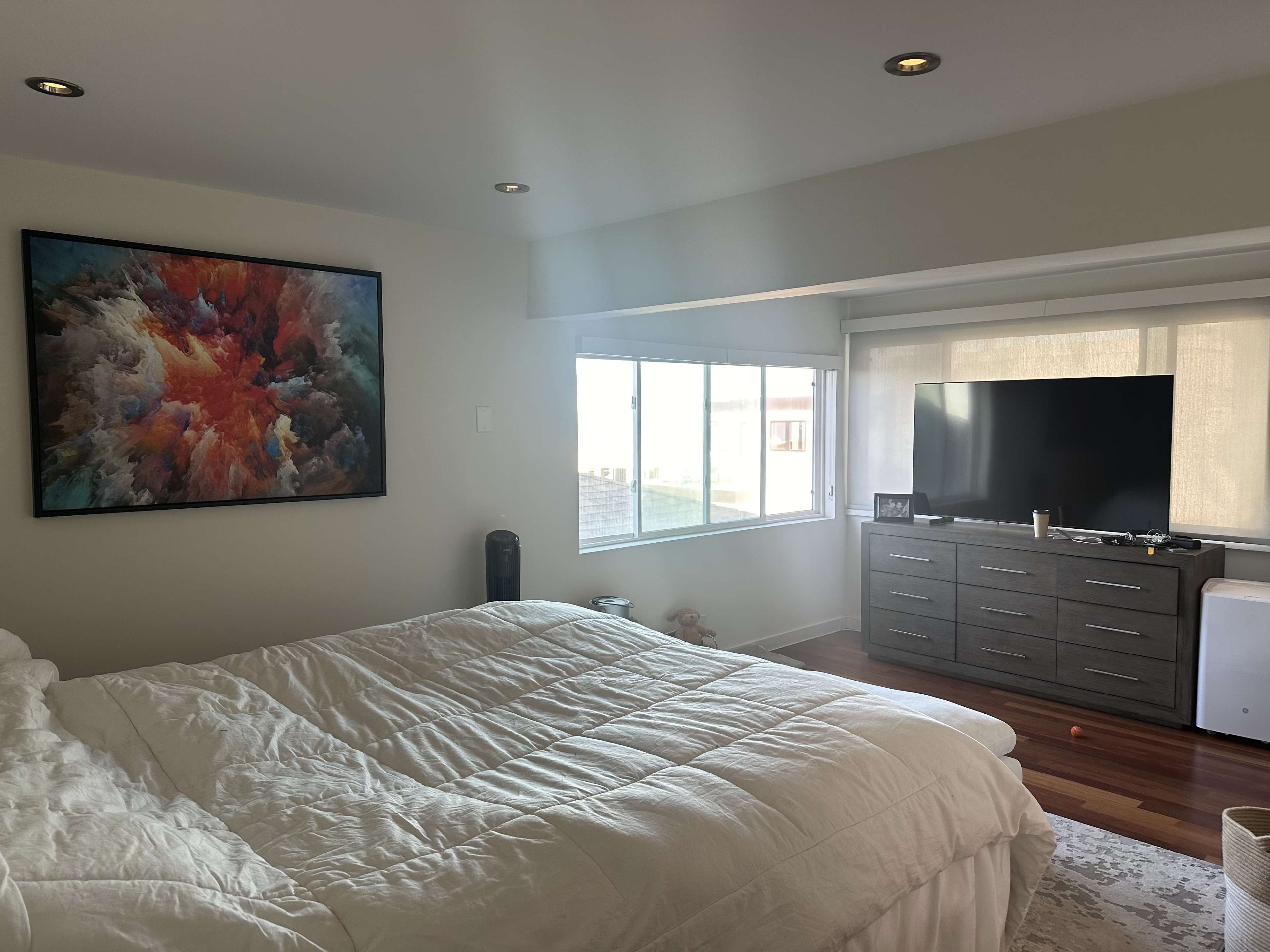 The image shows a well-lit bedroom featuring a large bed with a white comforter, a colorful abstract painting on the wall, a television on a dresser, and a window with sheer curtains.