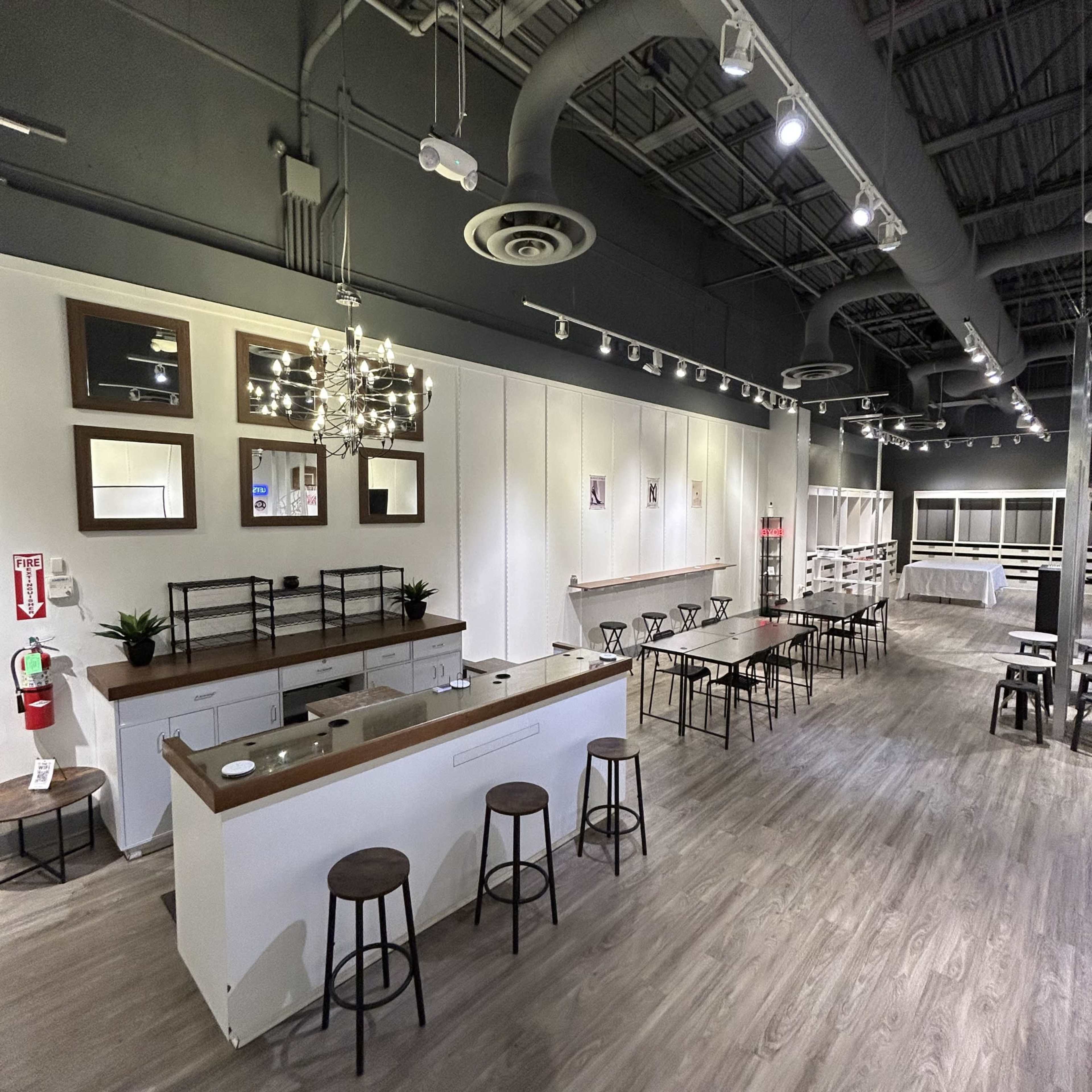 The image shows a spacious, modern interior of a venue with a bar area, several tables, and a chandelier, all arranged in a well-lit environment.