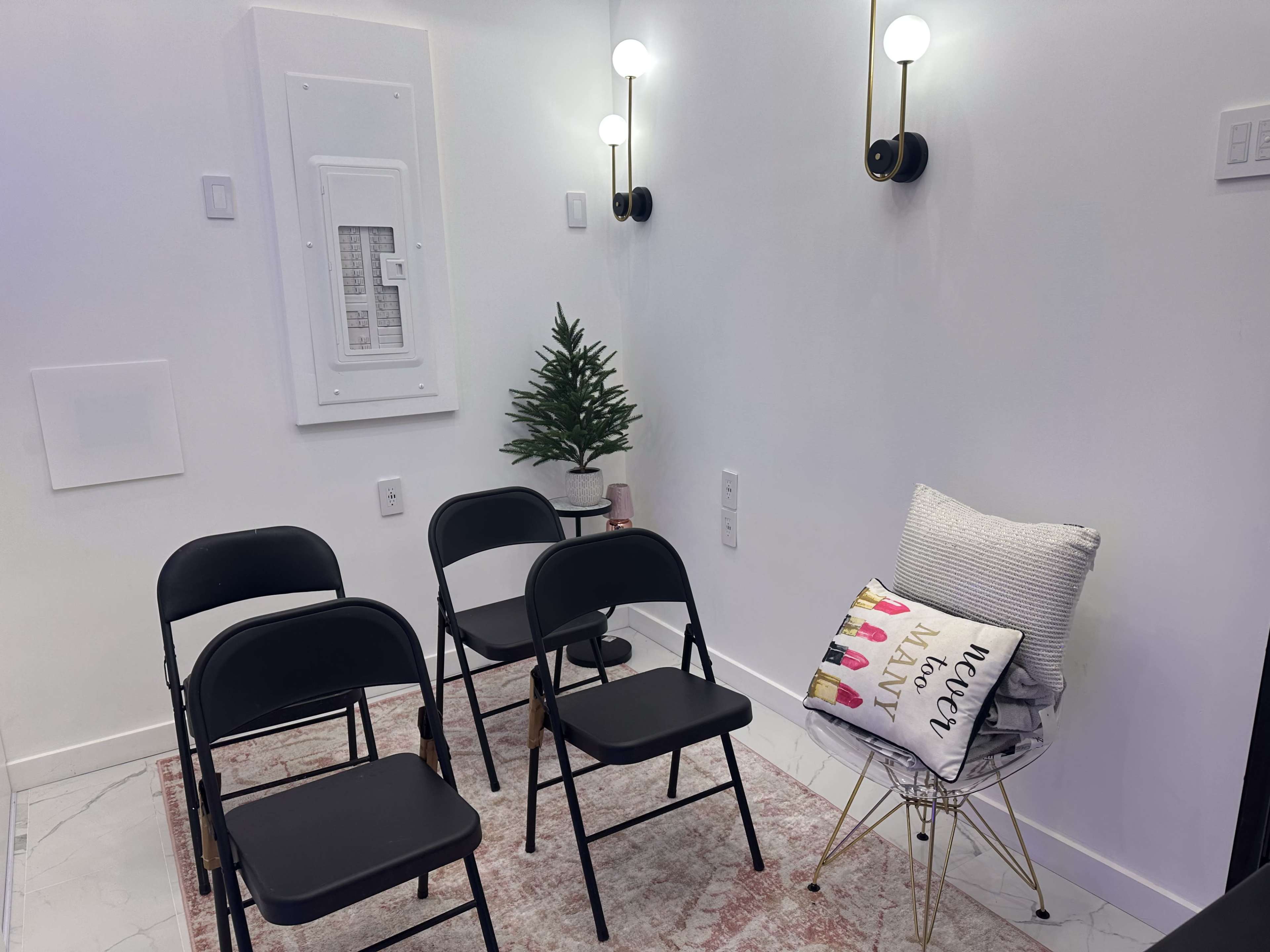 The image shows a small room with five black folding chairs arranged around a decorative rug, a small artificial plant in the corner, and a stack of pillows on a wire chair against a plain white wall.
