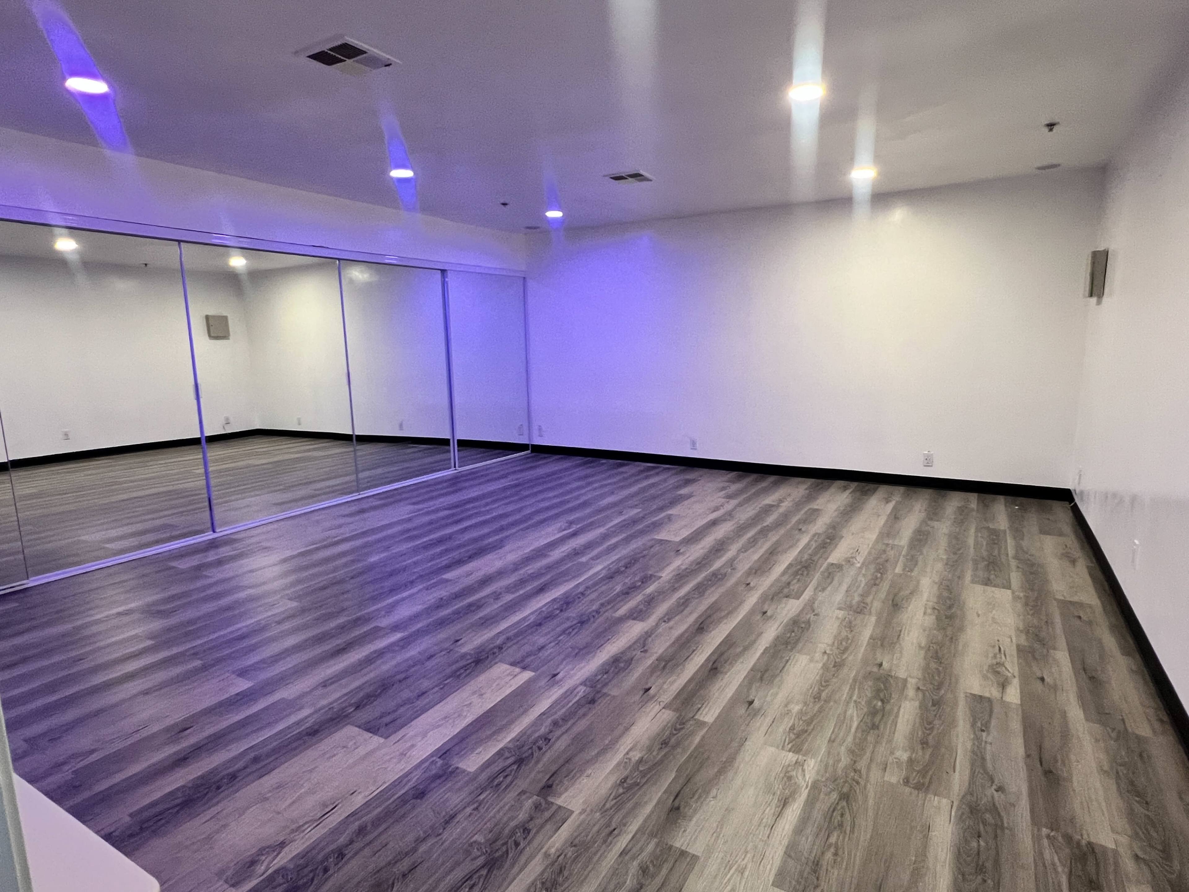 The image shows a spacious room with mirrored walls and wooden flooring, illuminated by soft purple lighting.