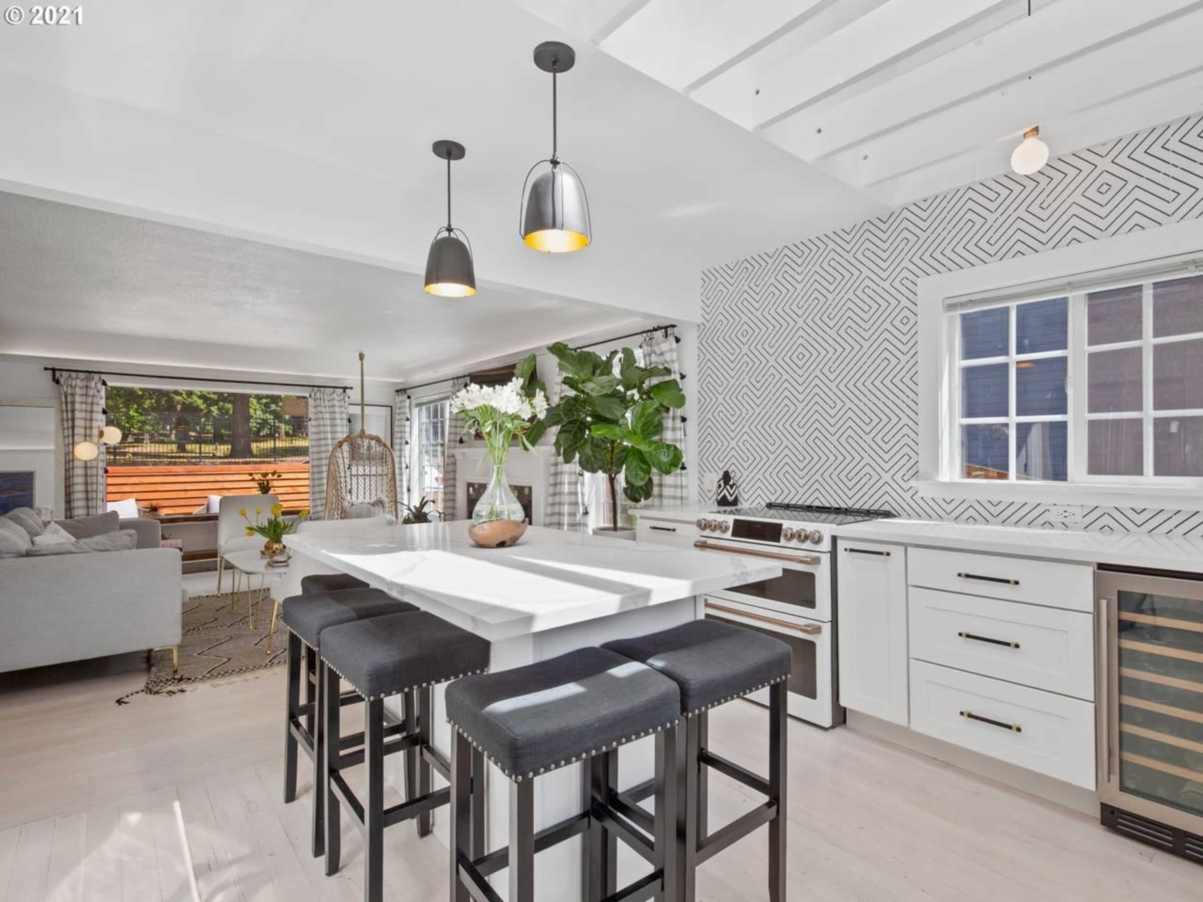 A modern kitchen with a large island, sleek appliances, and a patterned accent wall, featuring a view into a living area with a seating arrangement.