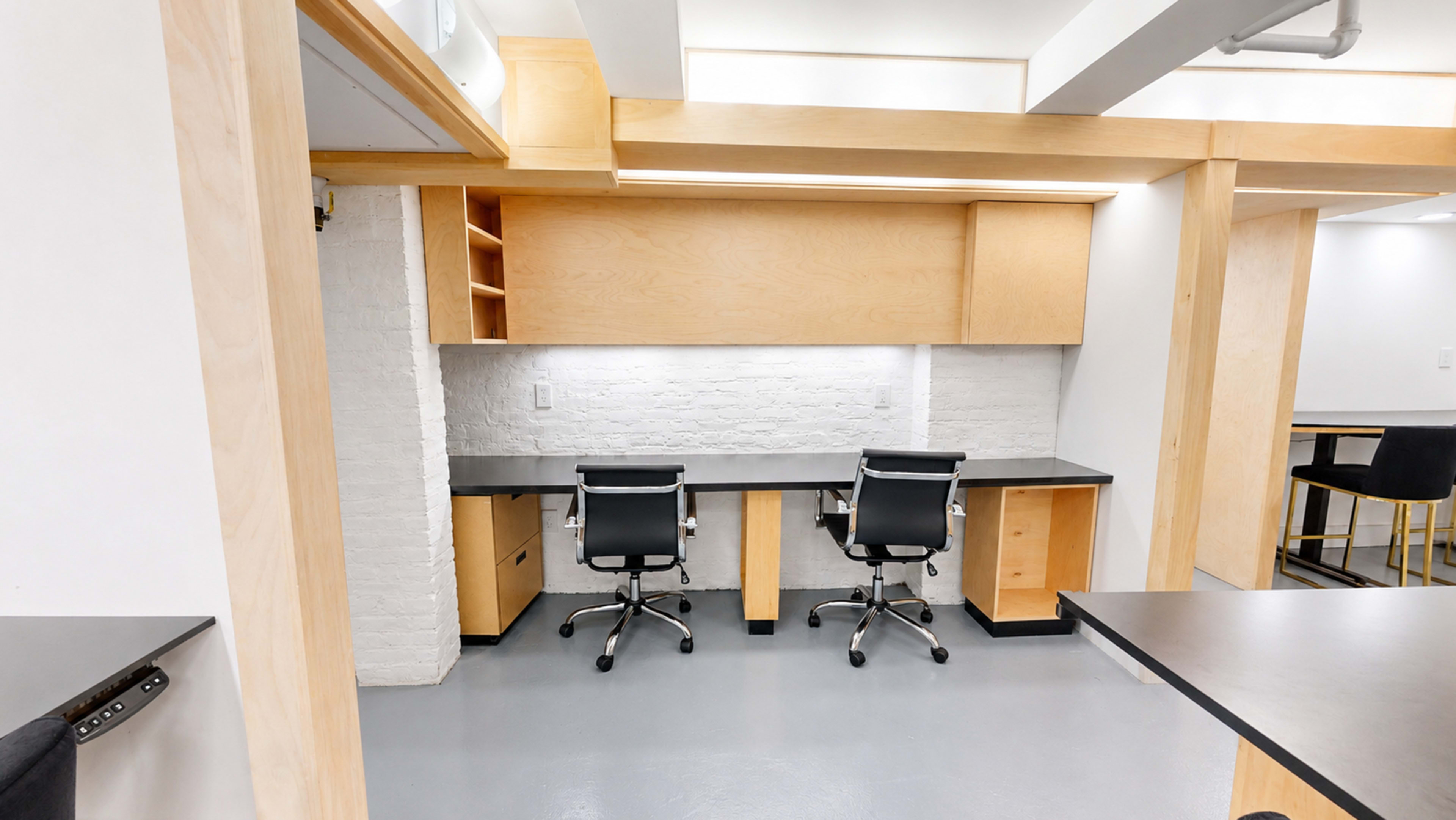 The image shows a modern workspace with two black chairs and a desk underneath a wooden storage unit against a white brick wall.