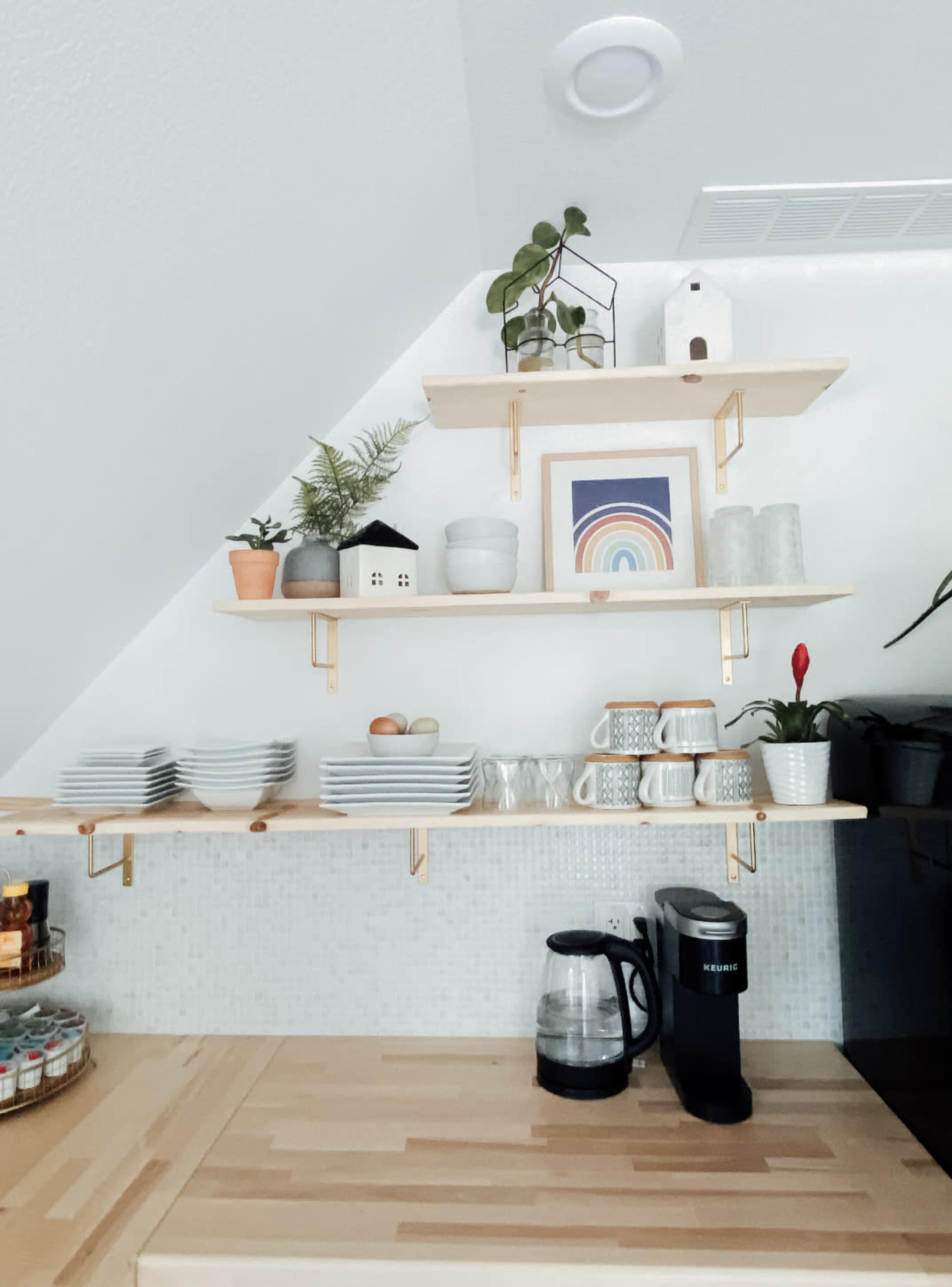 A set of wooden shelves mounted on a white wall, displaying dishes, decorative plants, a framed artwork, and a coffee maker on a wooden countertop.