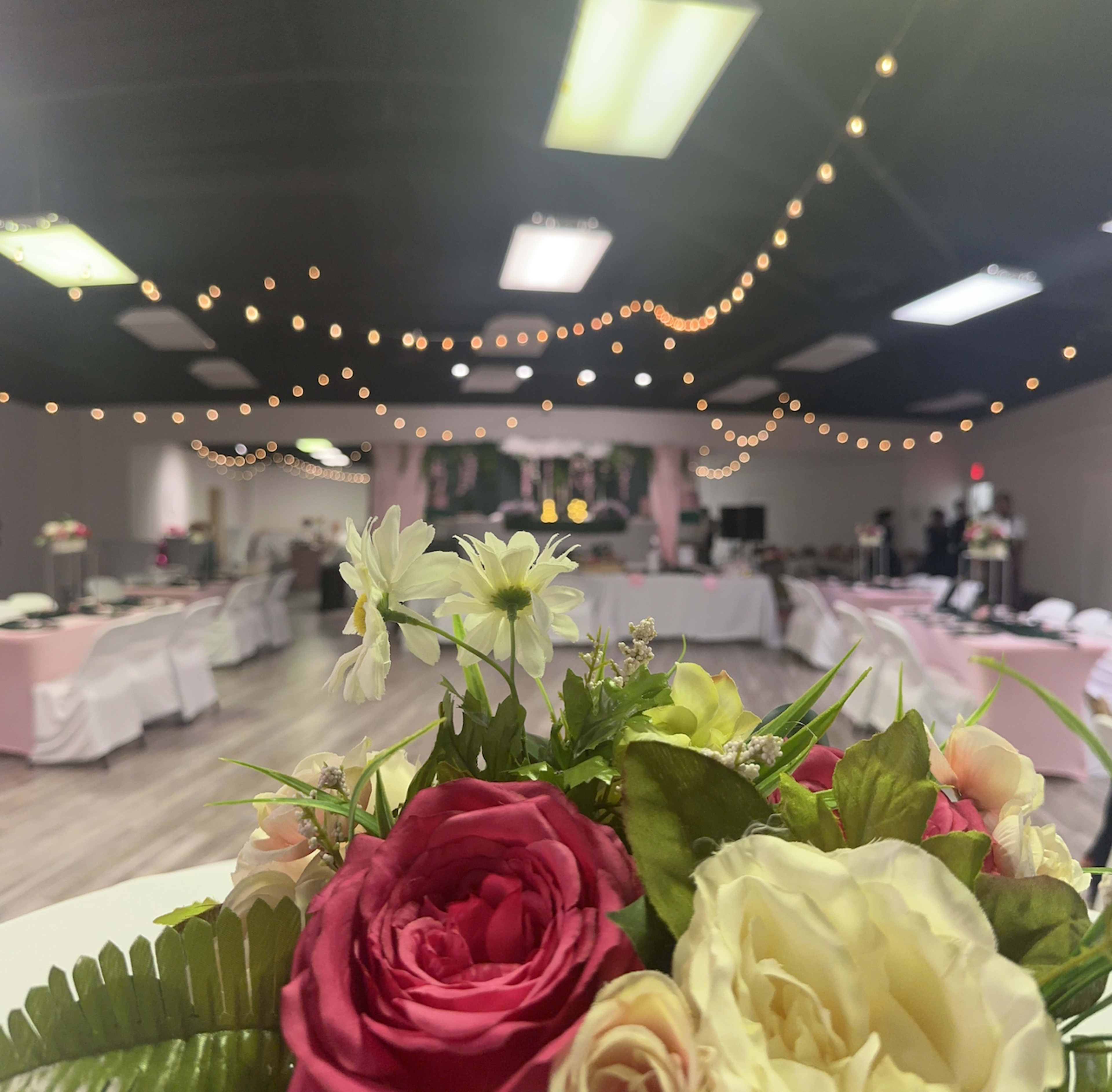 A floral centerpiece is prominently displayed in the foreground, while a decorated event space with tables and string lights is visible in the background.