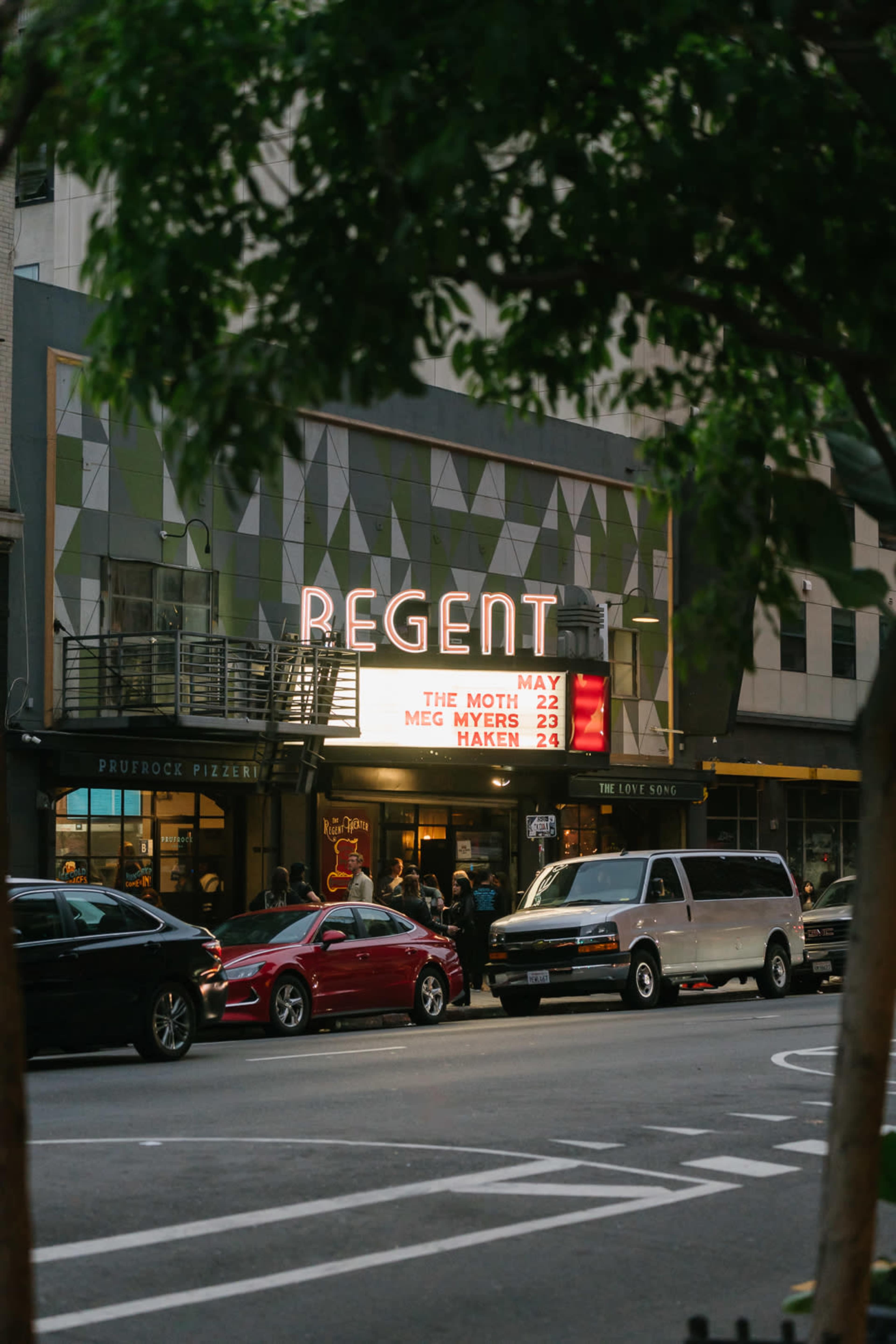 The Regent theater marquee displaying upcoming events, with a mix of parked cars and pedestrians in front.