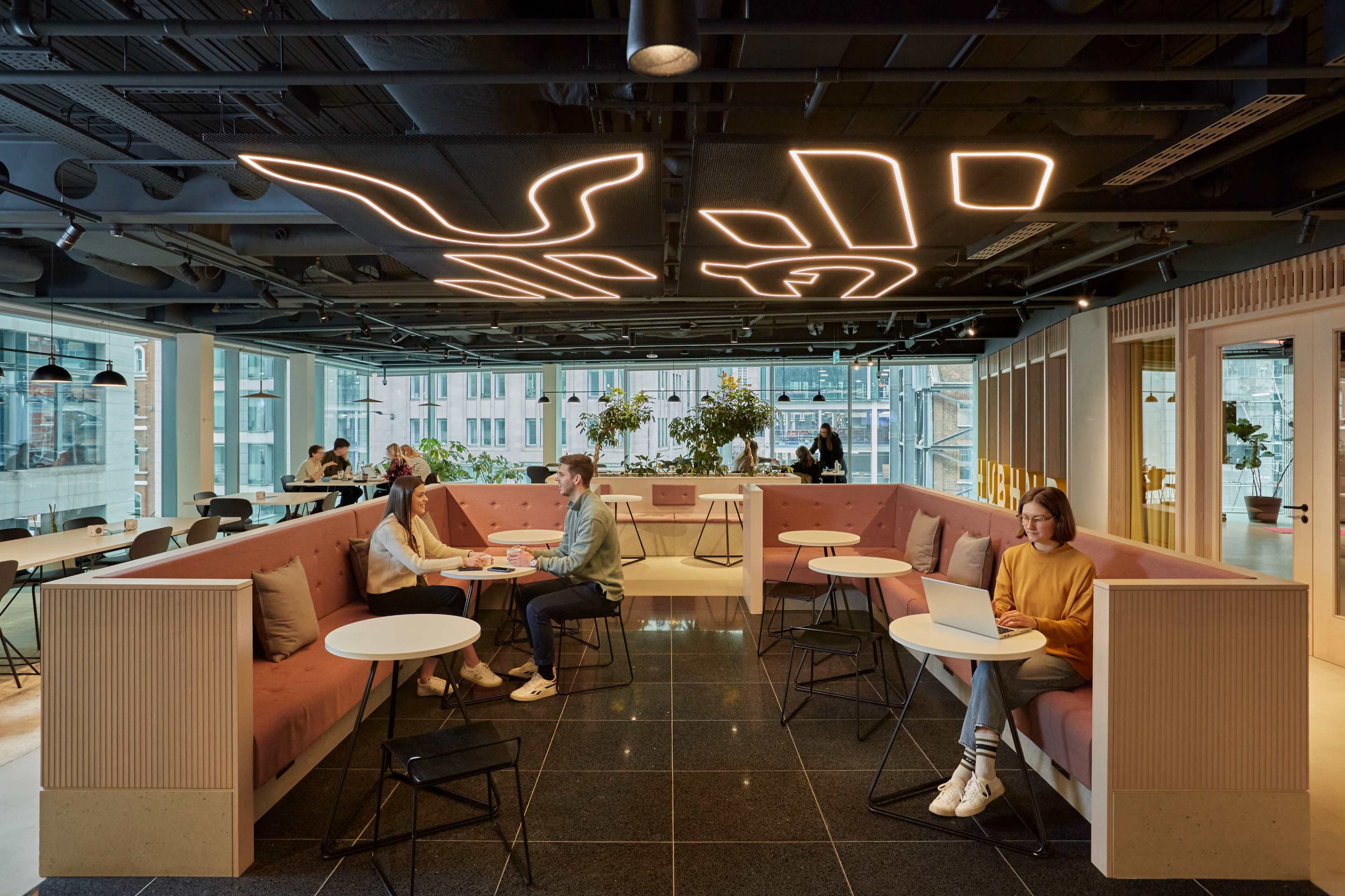 The image shows a modern café with pink seating areas, small tables, and people engaged in conversation and working on laptops.