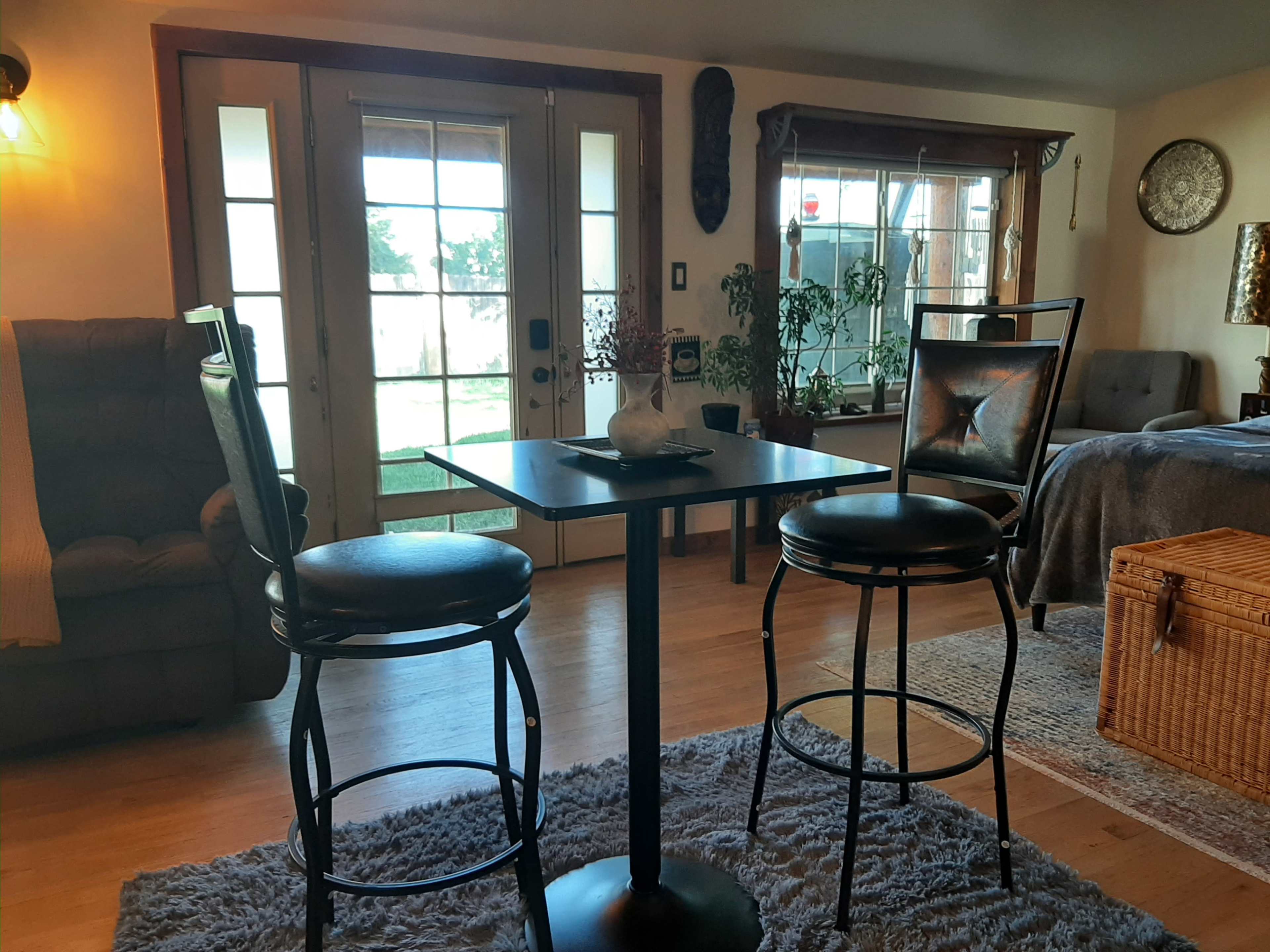 A small black table with two bar stools is positioned in a cozy living area with large windows and various plants.