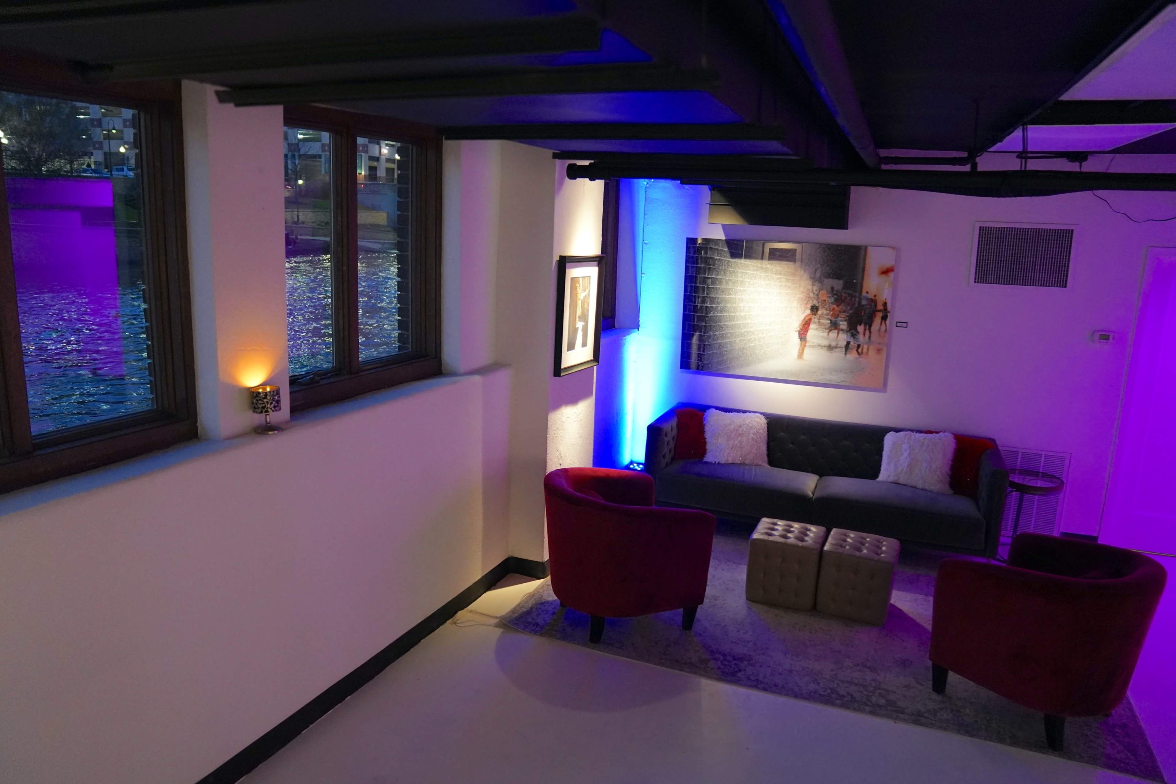 A modern lounge area with a dark sofa, two red chairs, and blue ambient lighting beside large windows overlooking a body of water.