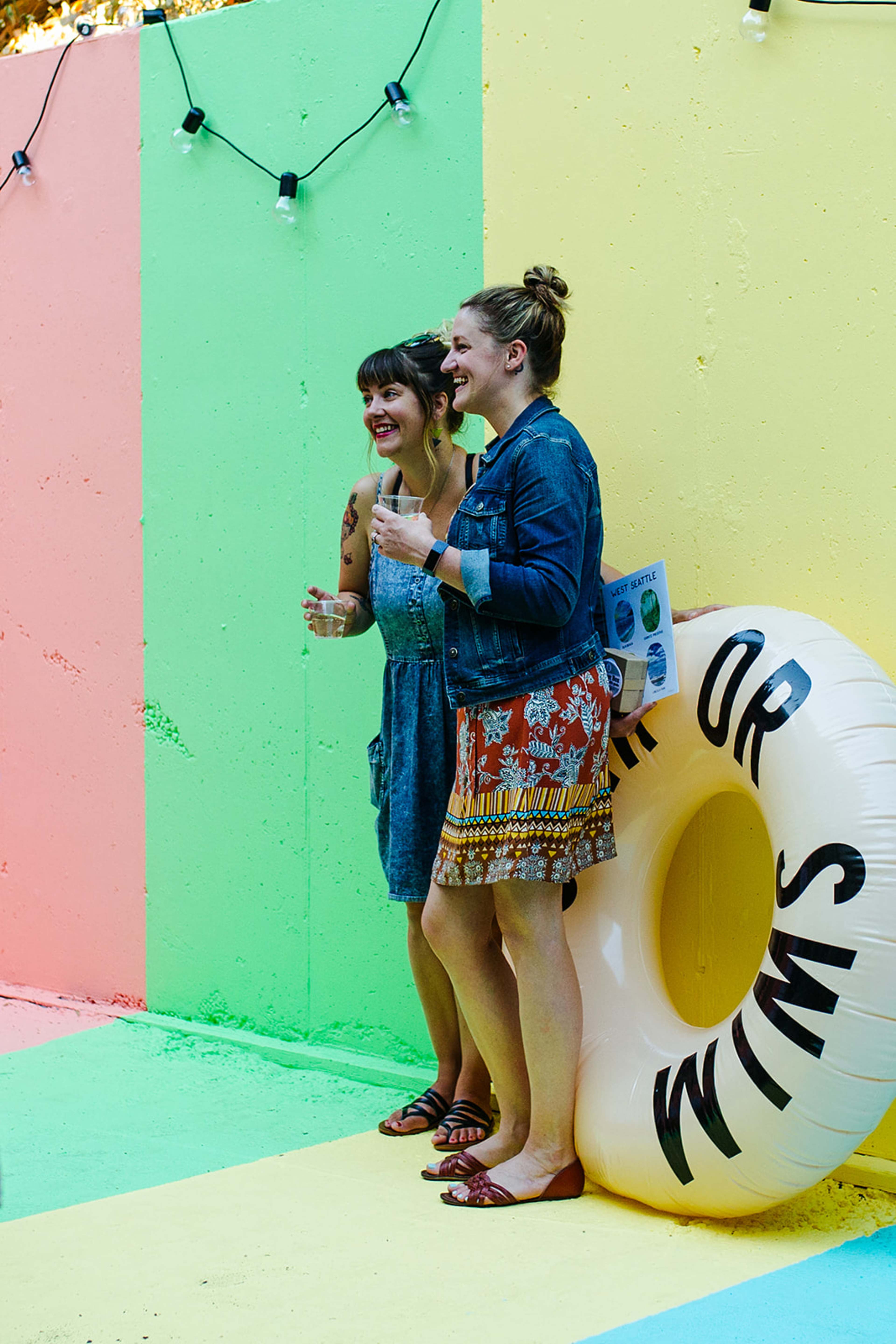 Two women stand beside a large inflatable pool ring, smiling and holding drinks in front of a colorful wall.