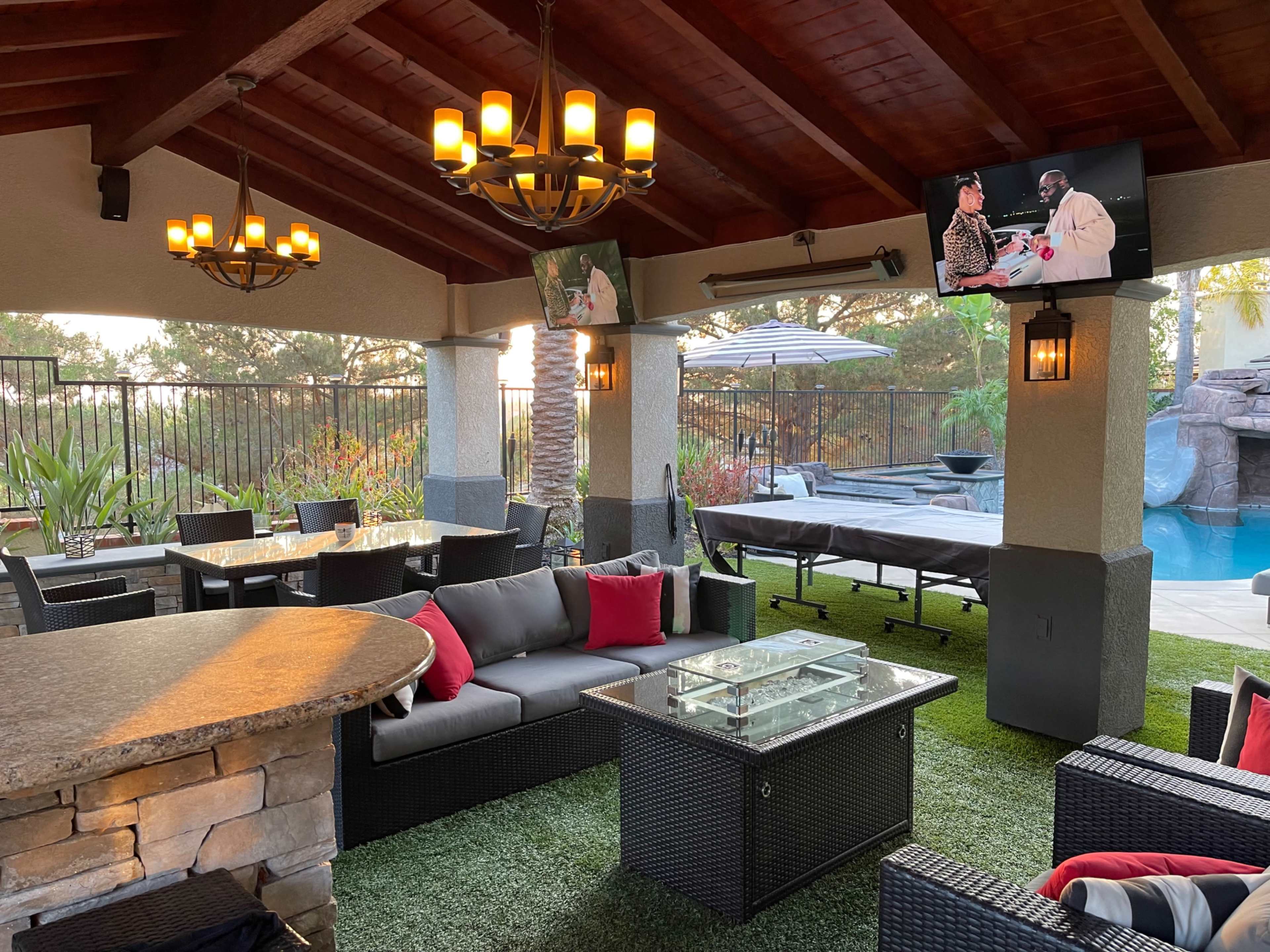 The image shows a covered outdoor lounge area with comfortable seating, a stone table, and a television, overlooking a pool and landscaped surroundings.