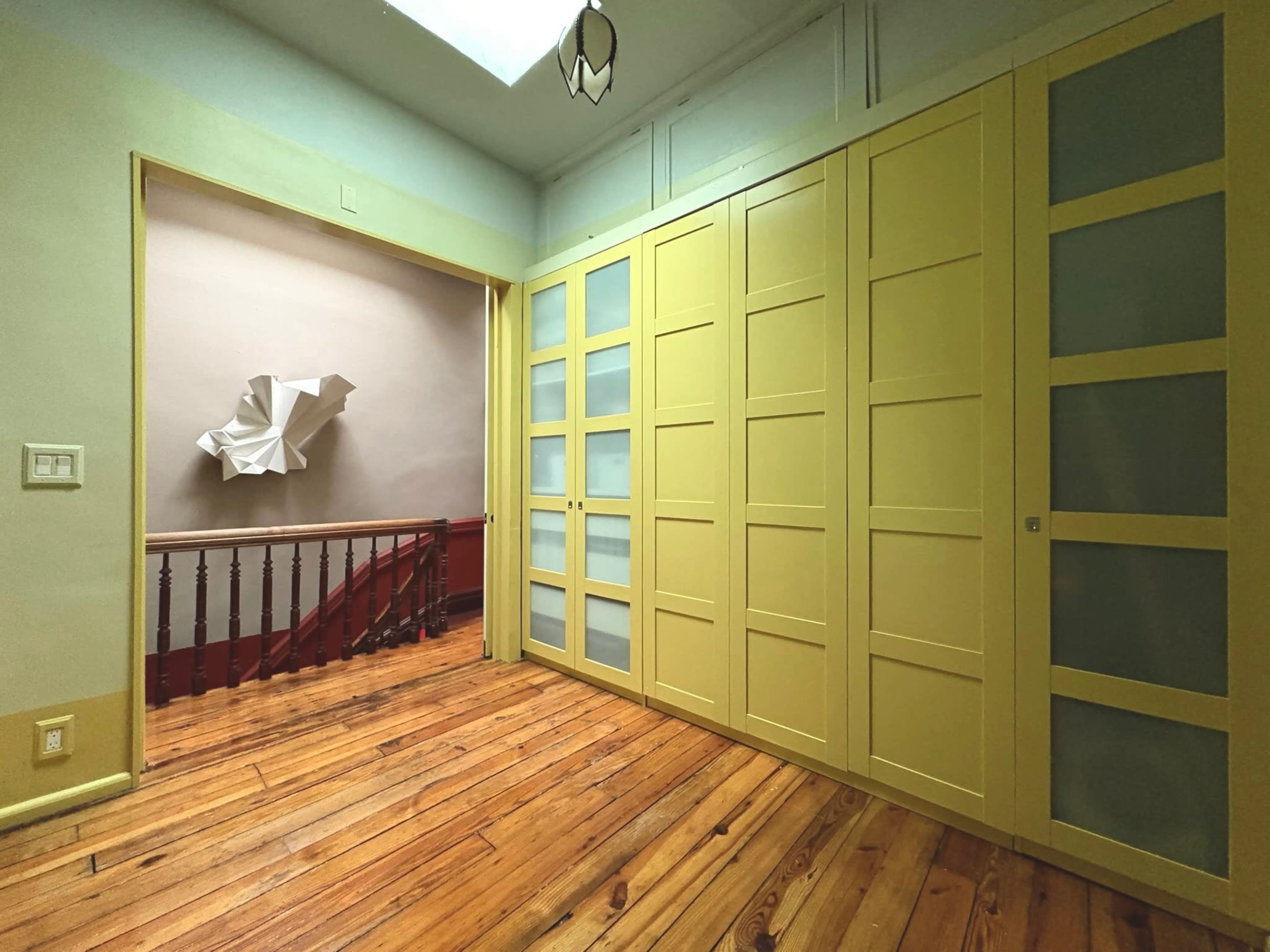 The image shows a room with yellow cabinetry, hardwood flooring, and a sculptural artwork on the wall, with a staircase visible through an open doorway.
