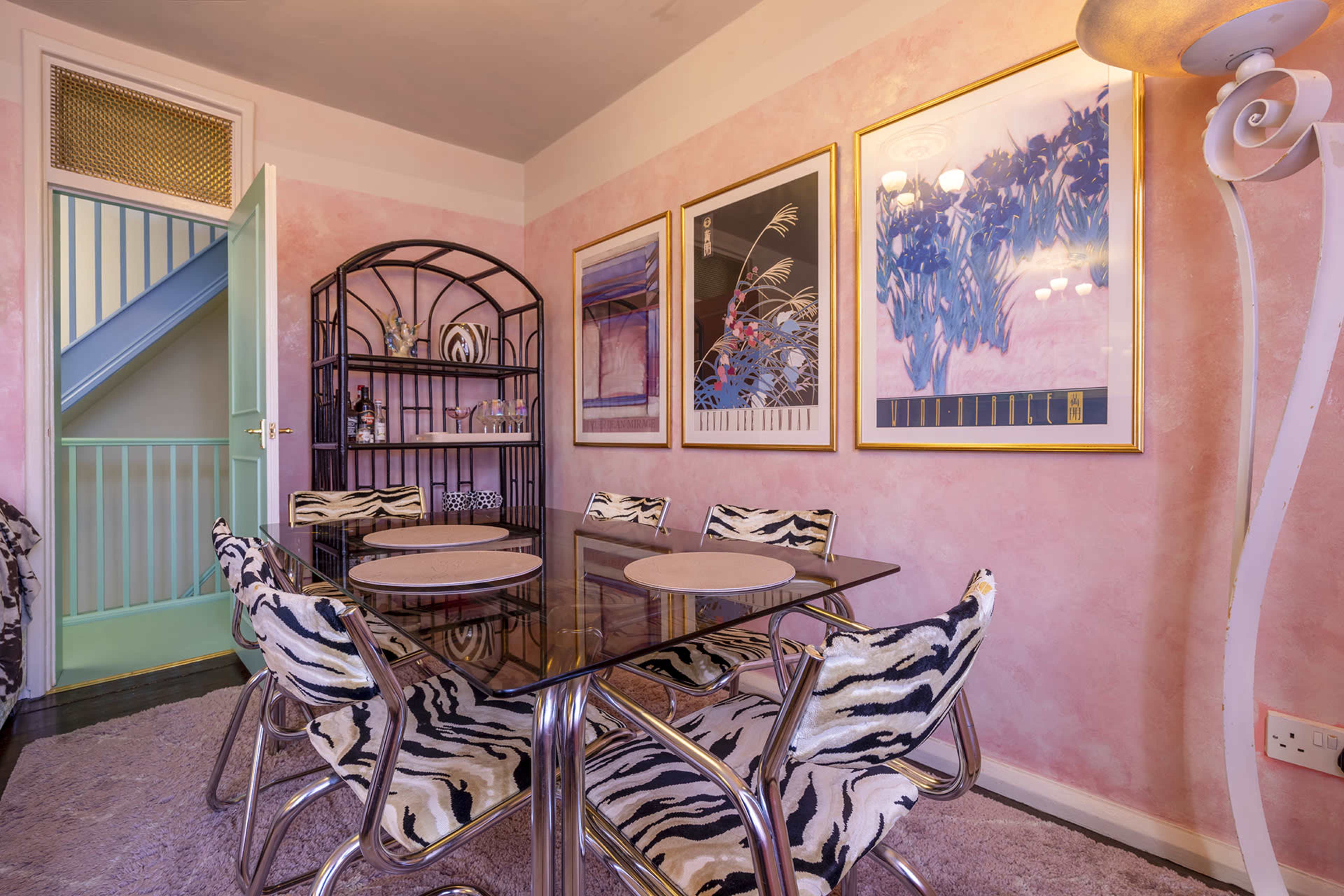 A dining area features a glass table surrounded by zebra-striped chairs, with pink walls and framed artworks, complemented by a turquoise door leading to another space.