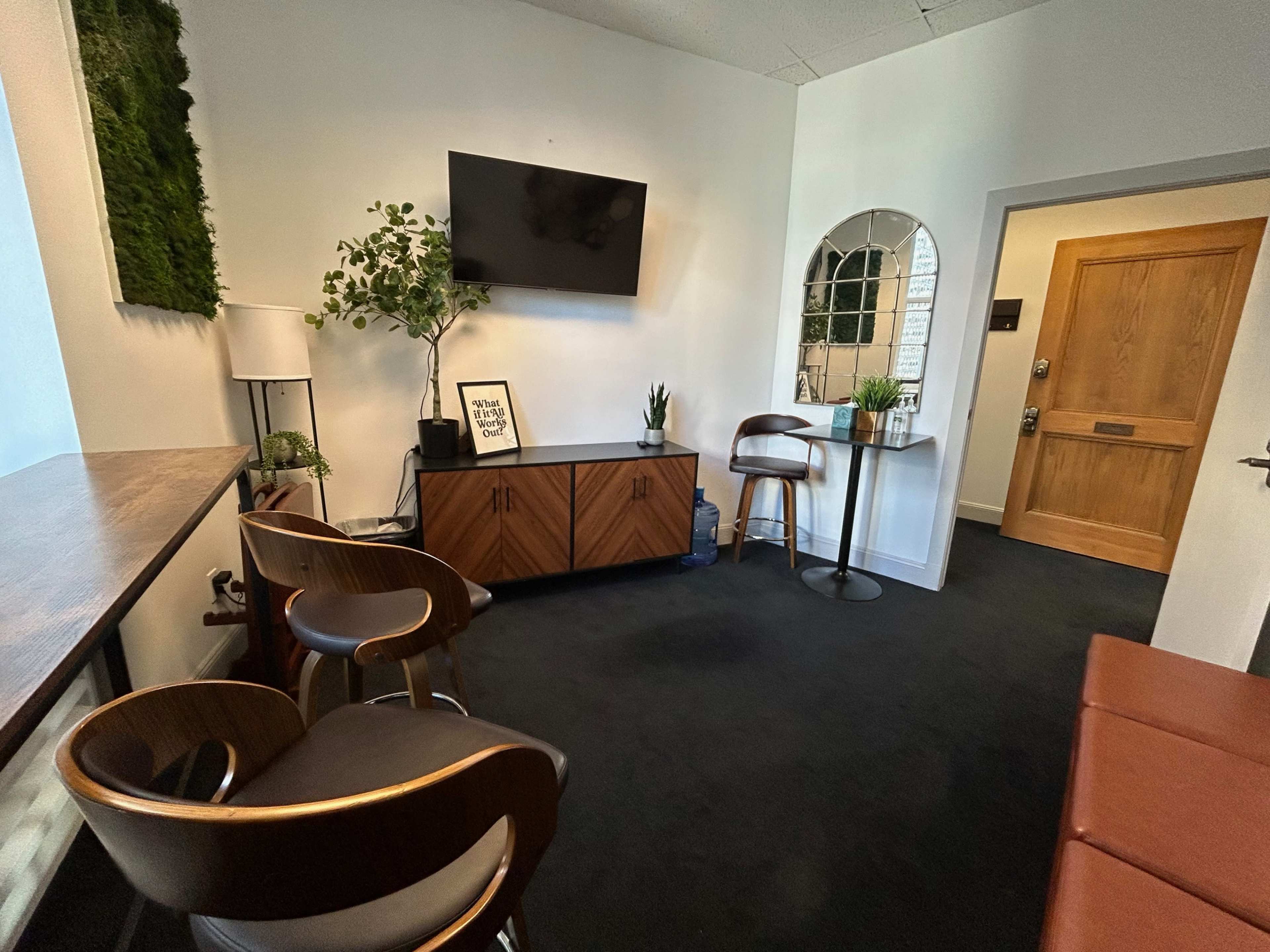 A cozy waiting area with a wall-mounted TV, plants, and modern furniture, including two chairs and a small table.