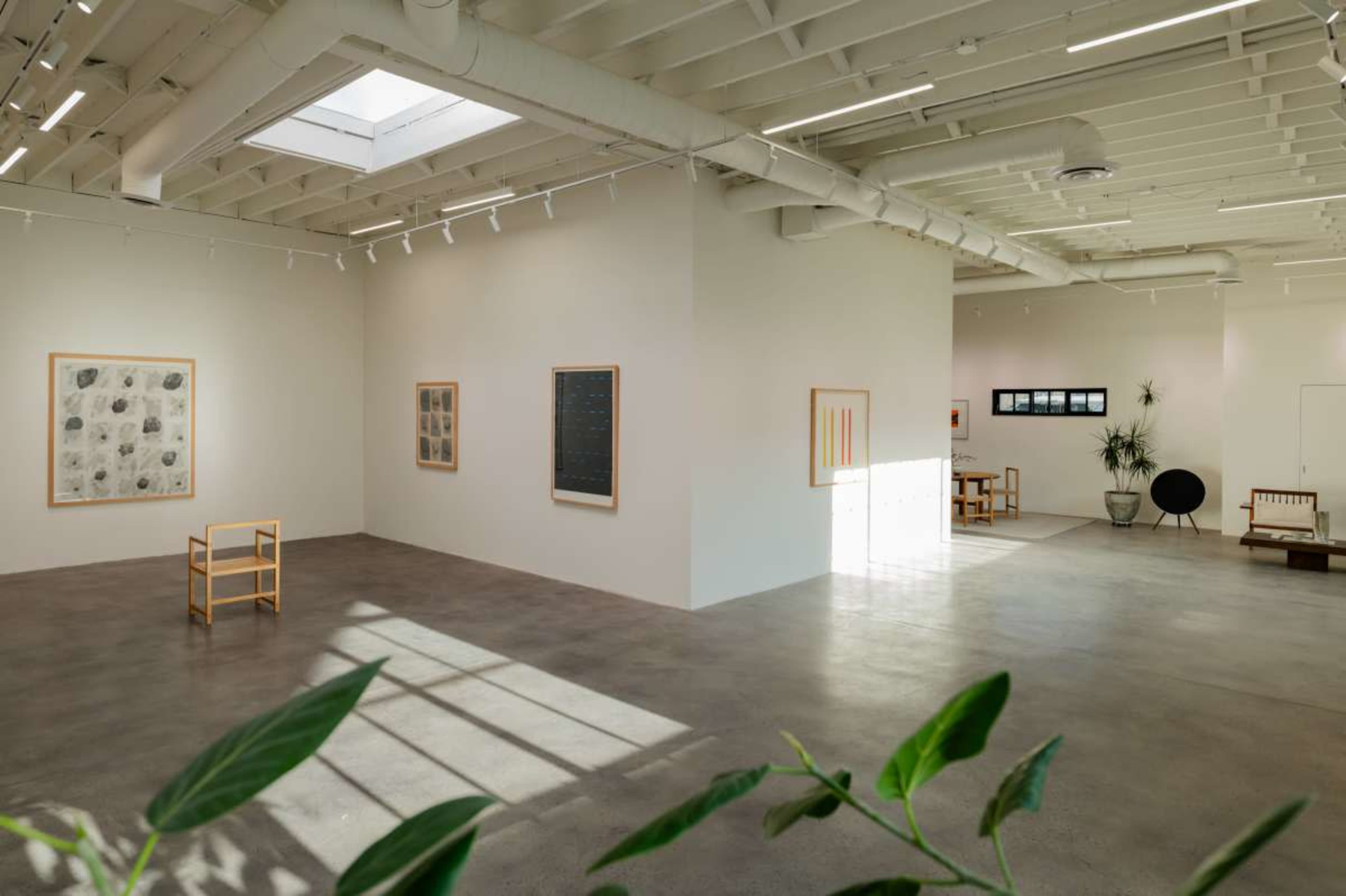 The image shows a minimalist art gallery featuring light-colored walls, various artworks framed on the walls, and a few pieces of furniture scattered throughout the space.