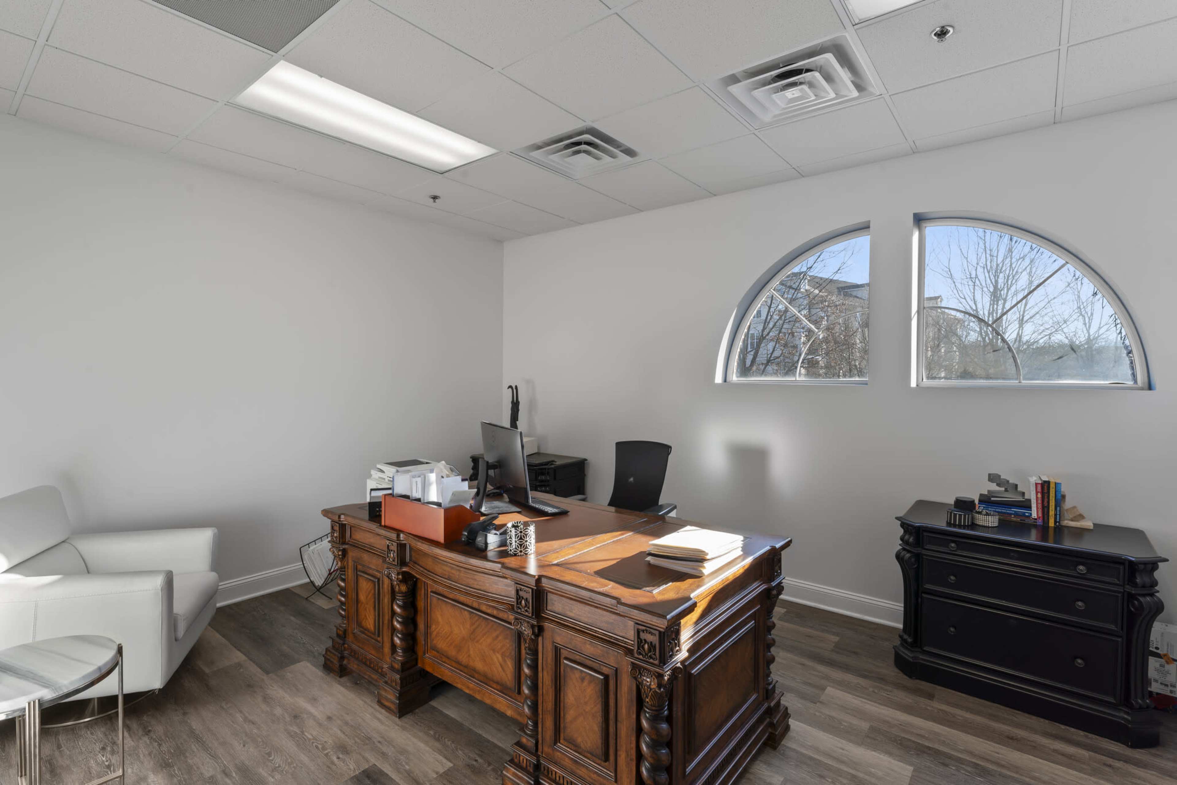 The image shows a modern office space featuring a large wooden desk, a chair, and two arched windows that allow natural light to enter.