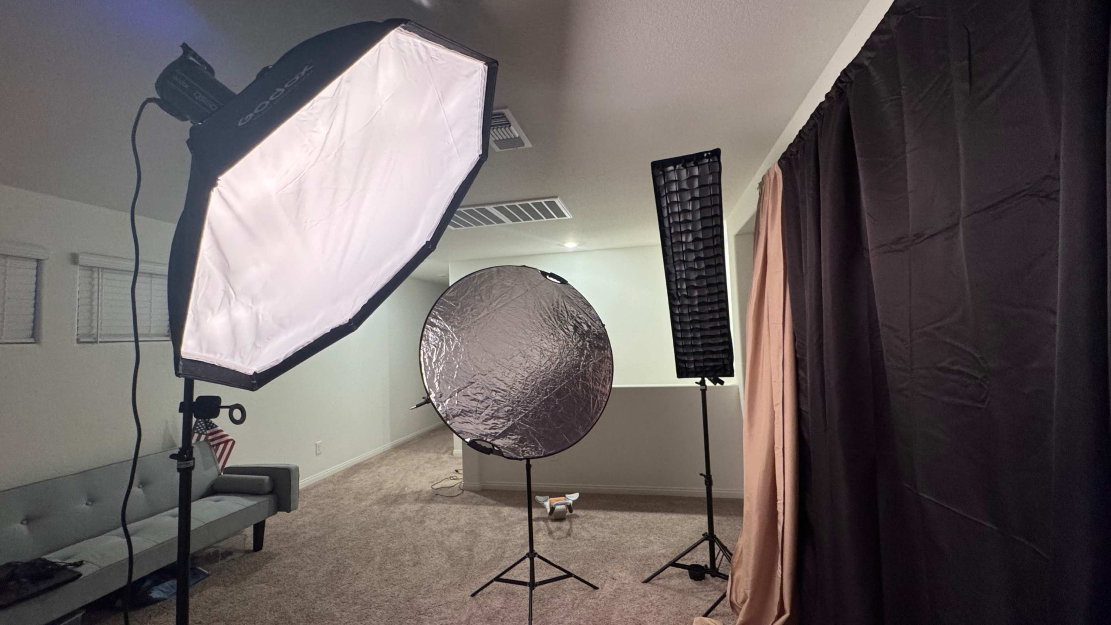 The image shows a photography setup in a room, featuring softbox lighting, a reflector, and a background curtain, with a sofa visible in the background.