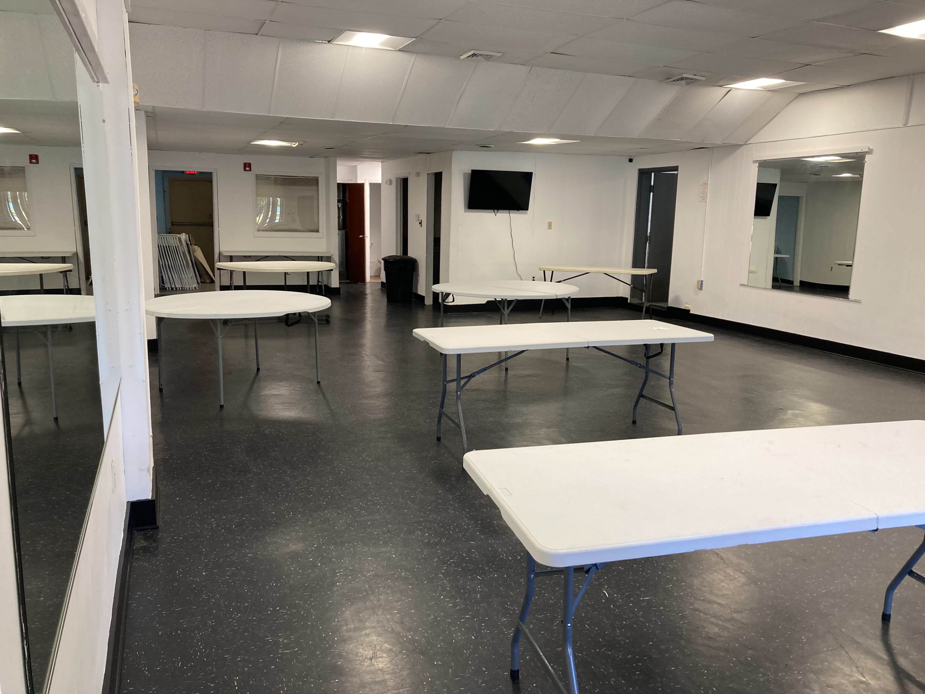 A large, empty room with several plain white tables arranged throughout, two mirrors on one wall, and a television mounted on another.