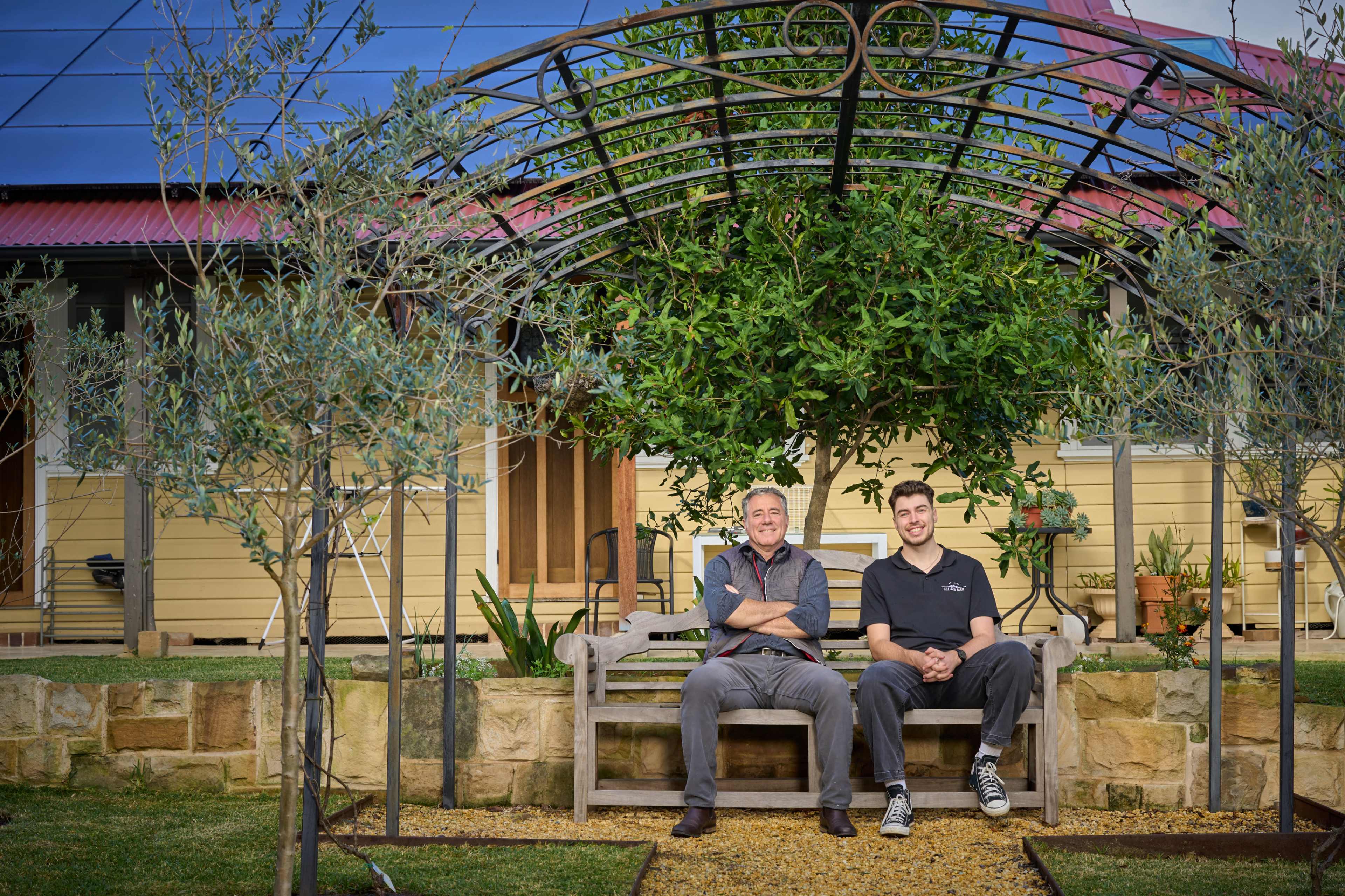 Two men sit on a wooden bench under a trellis adorned with greenery in a garden setting, with a house visible in the background.