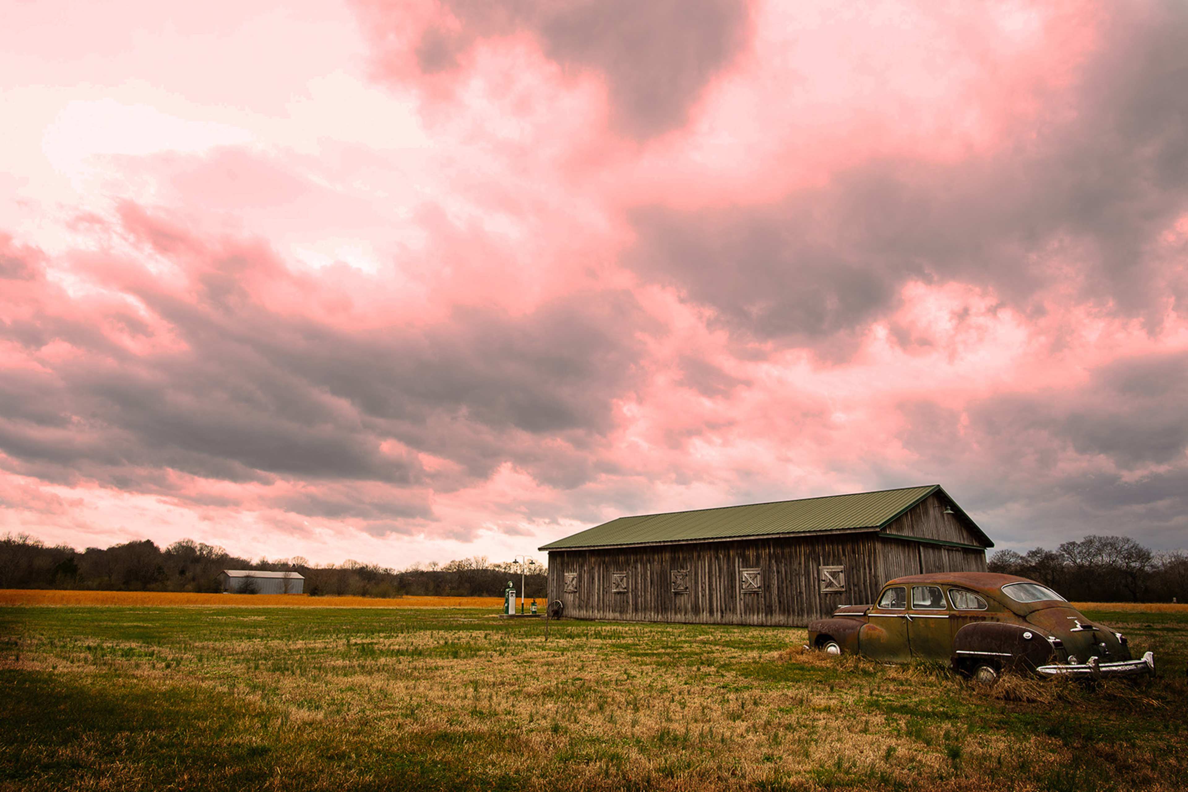 A weathered barn stands beside an old car in a grassy field under a dramatic pink and gray sky.