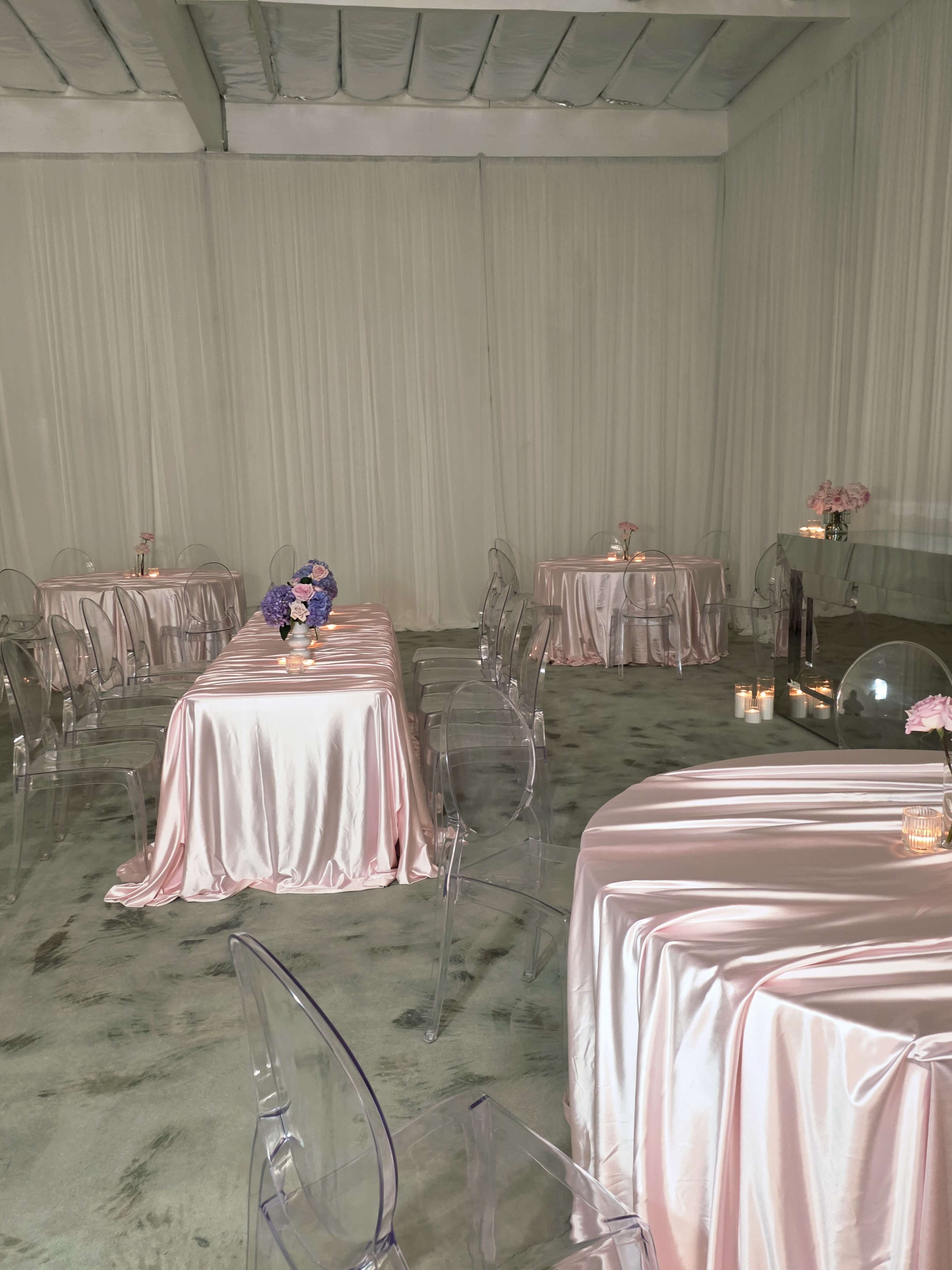The room features several tables covered with pink satin linens and transparent chairs, set against a backdrop of soft fabric.