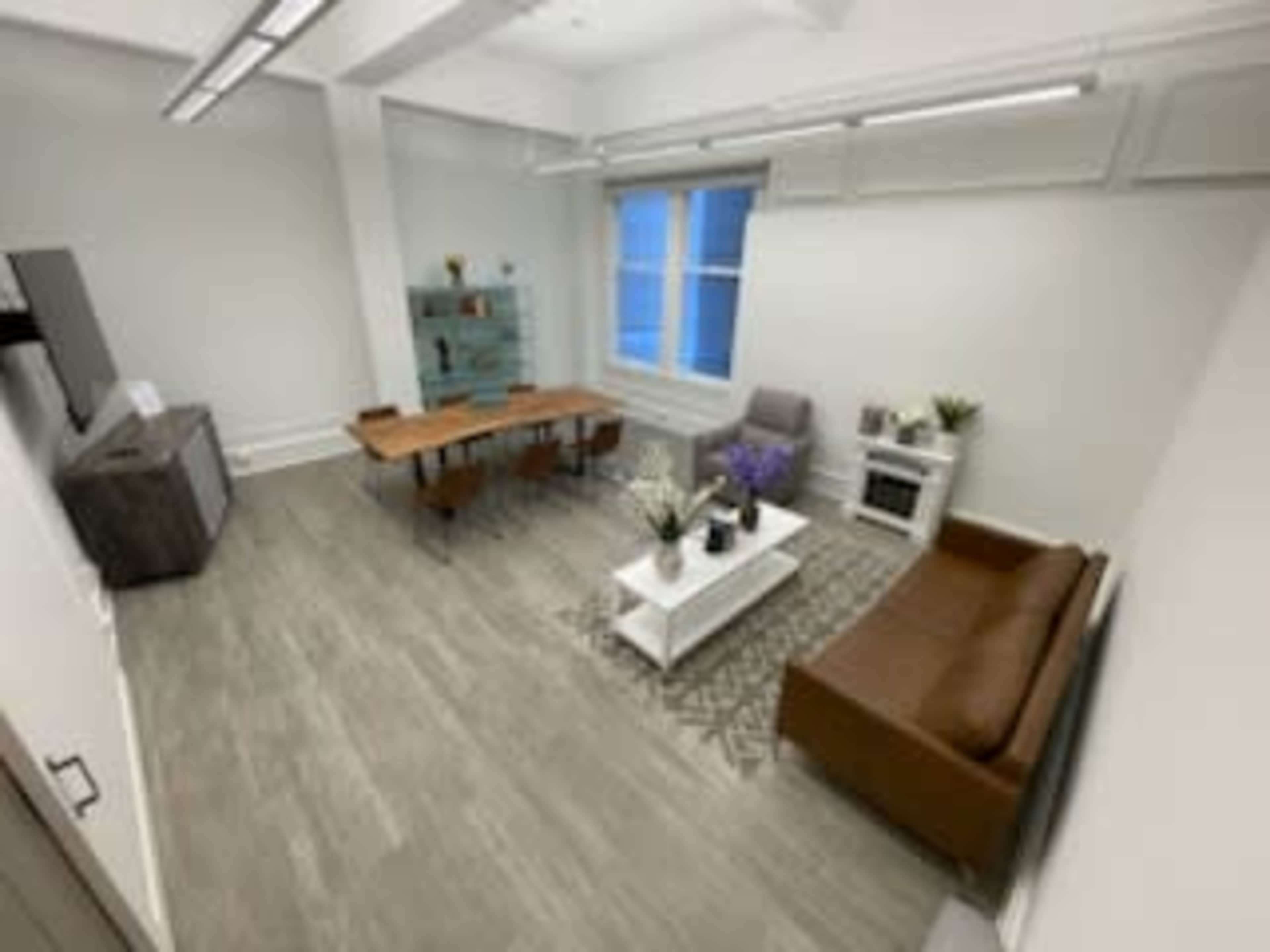 The image shows a spacious, modern office with a wooden table, a sofa, a coffee table, and a bookshelf, all arranged in a well-lit room.