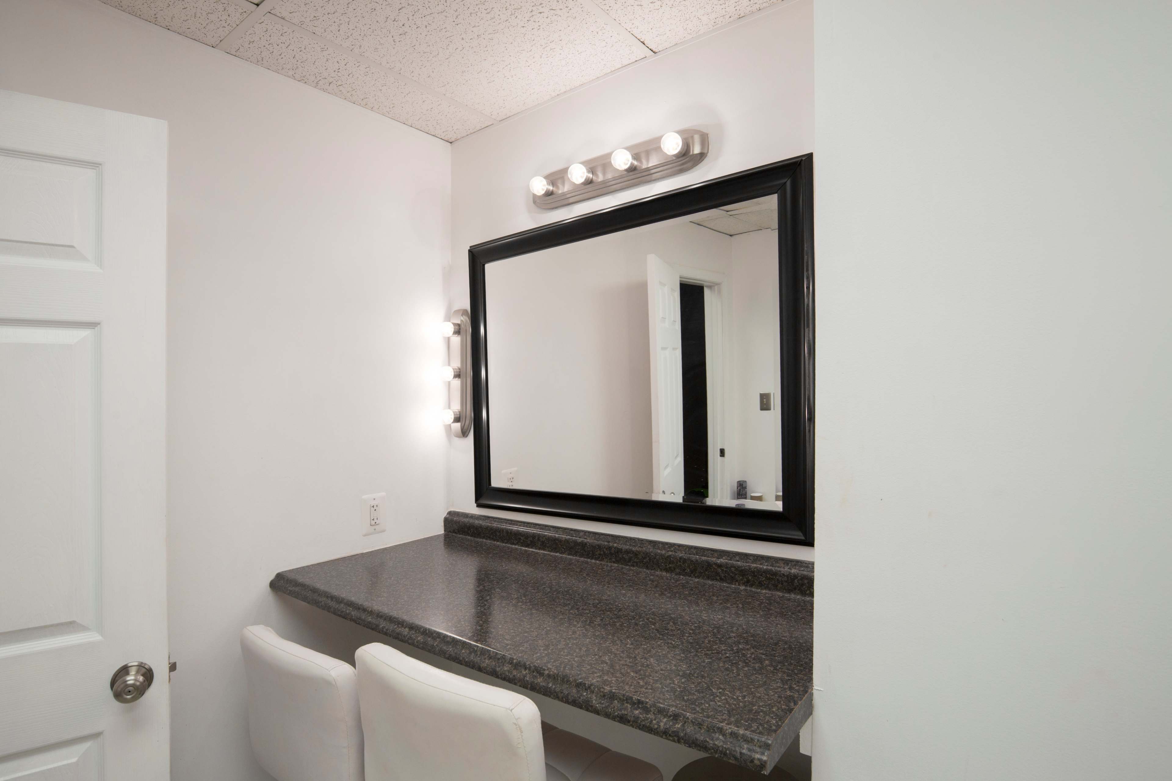 A clean, well-lit bathroom area features a large mirror above a dark countertop with two white chairs and a door leading to another room.
